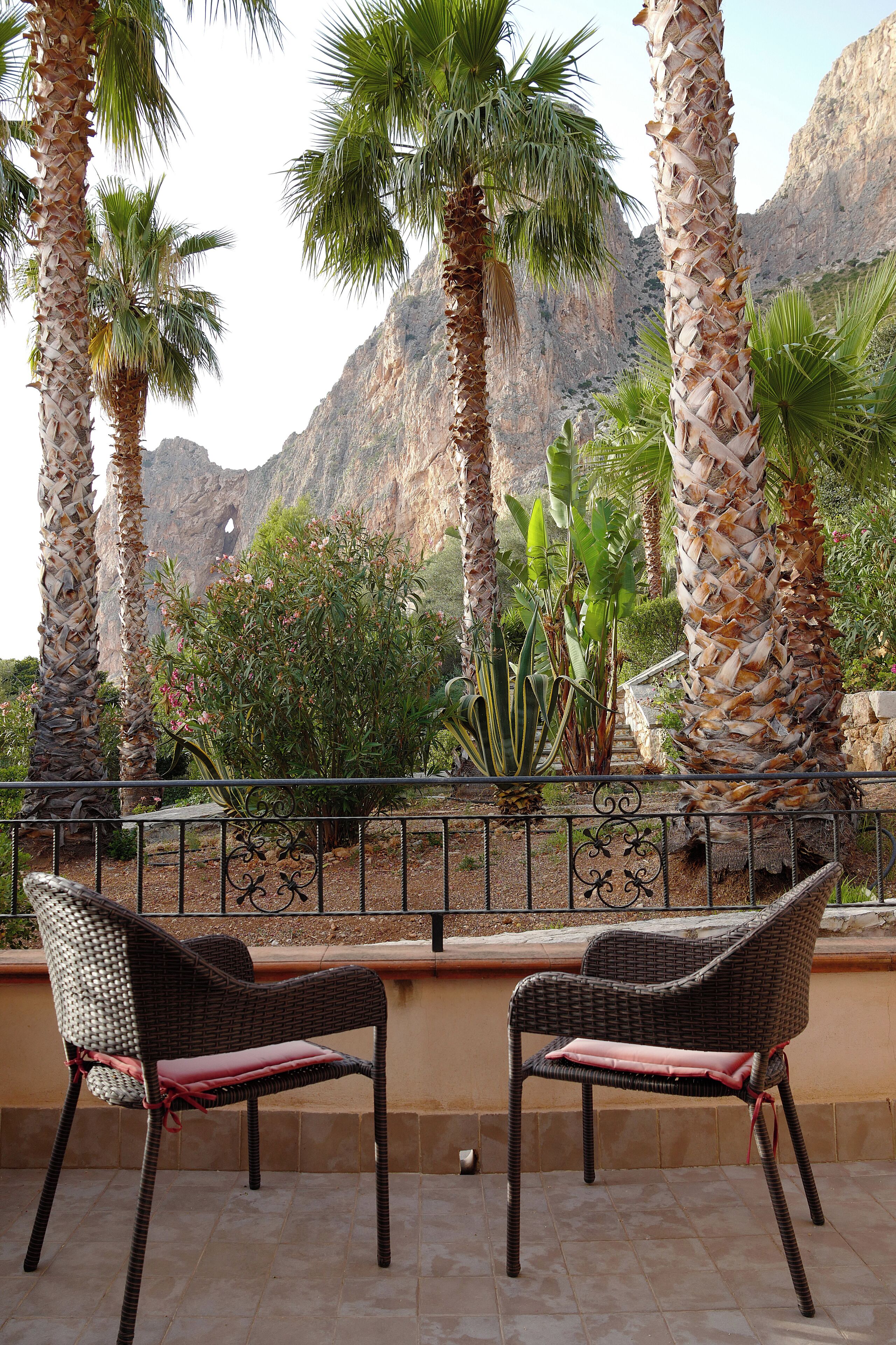 A terrace with wicker chairs and a view of palm trees and rocky mountains.