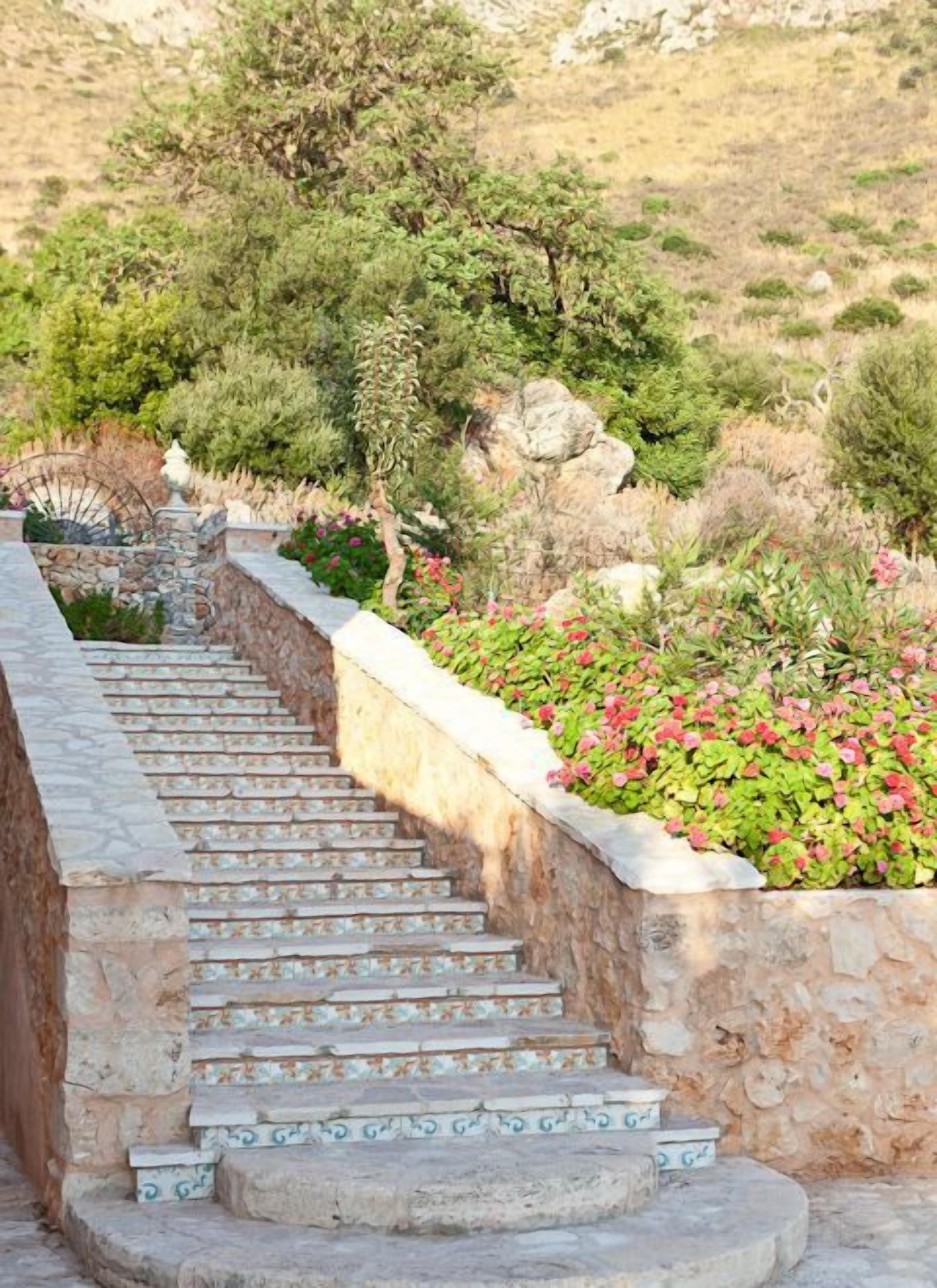 A stone staircase with patterned tiles leading up a hill, surrounded by greenery and flowers.