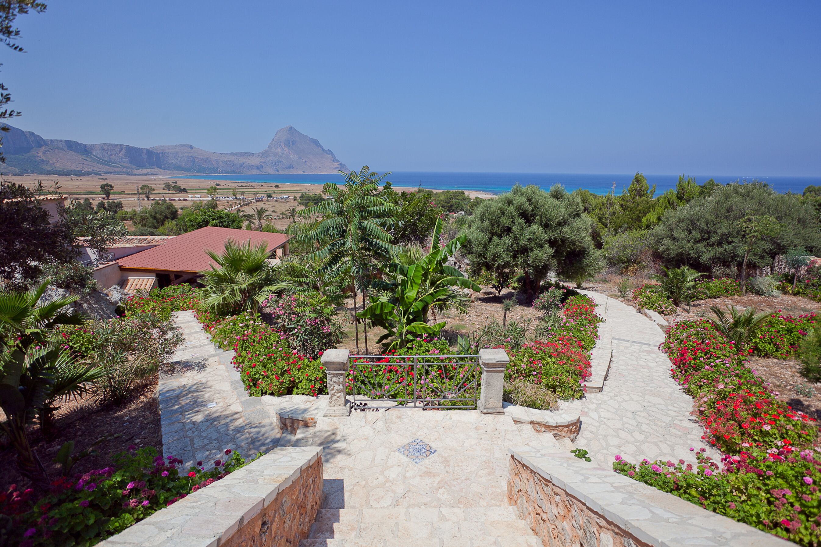 A garden pathway leading to a view of the sea, with a mountain in the background.