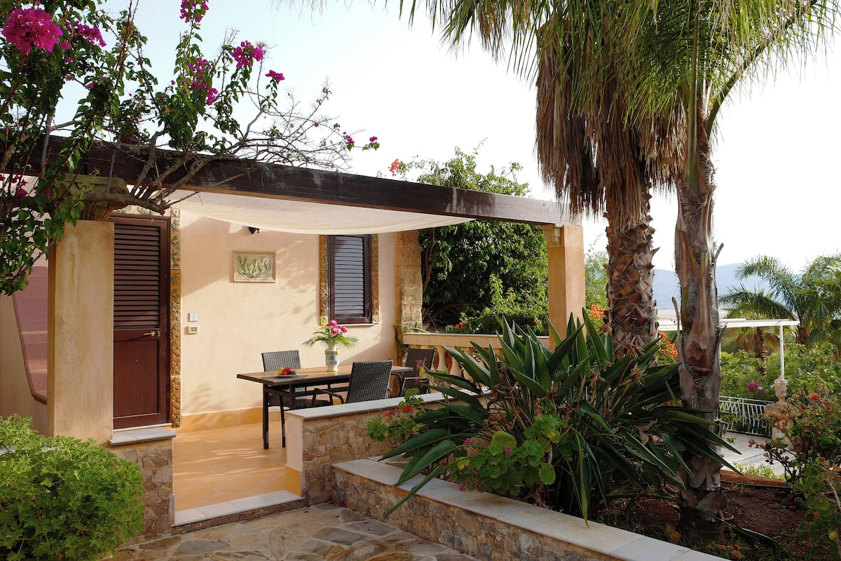 A covered outdoor seating area with a table and chairs, a door, and a stone wall.