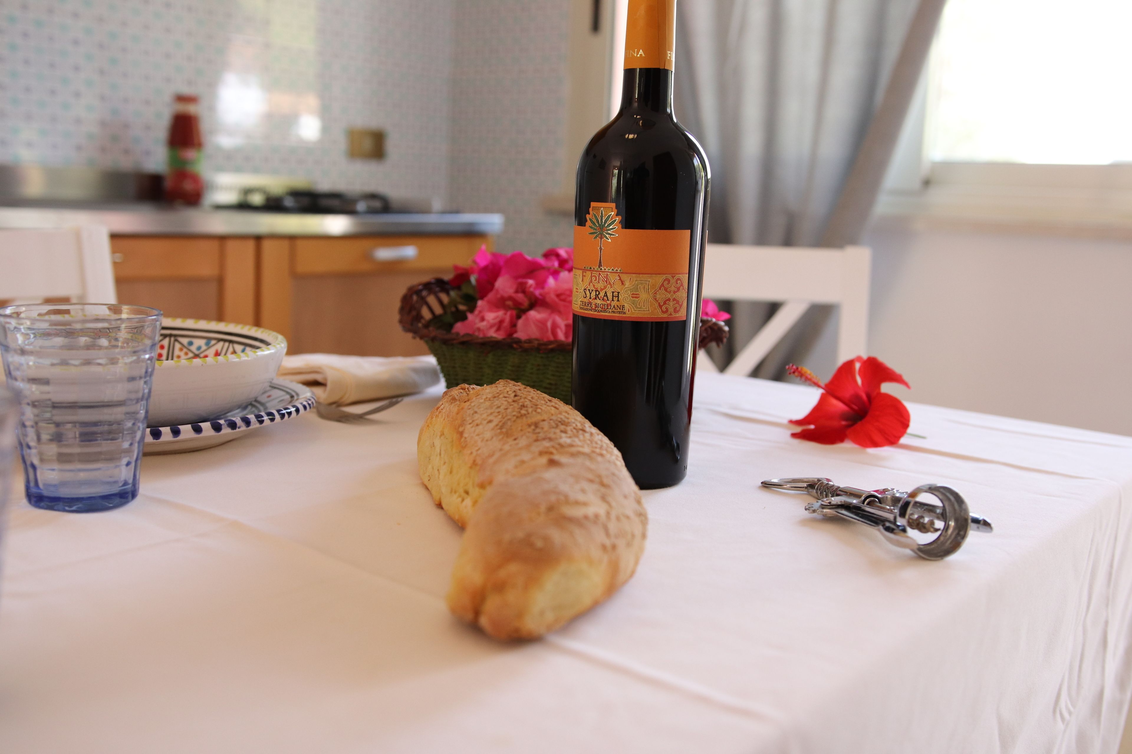 A table with a bottle of wine, a glass of water, a plate with a croissant, a bowl of flowers, and a red flower on a tablecloth.