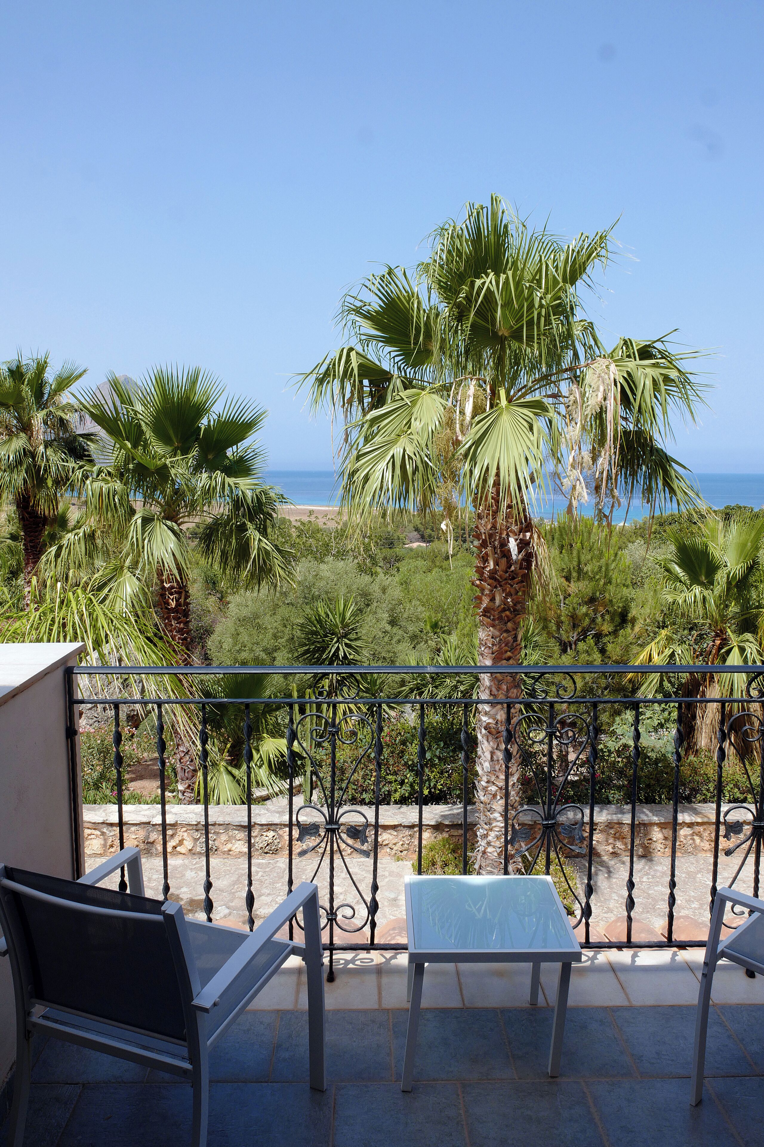 Un balcón con dos sillas y una mesa, con vista a una playa con palmeras y el océano.