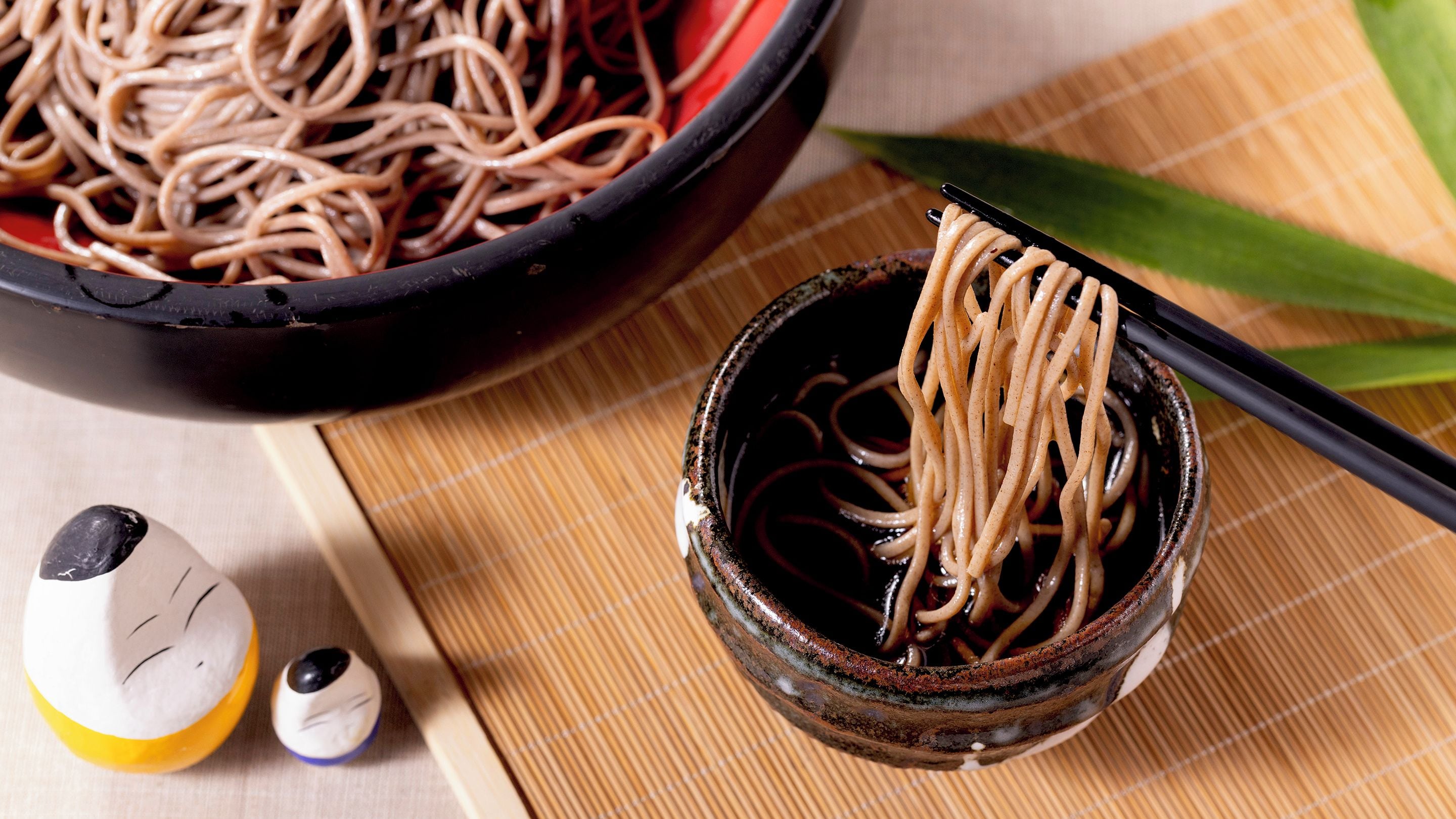 A bowl of noodles with chopsticks, a small bowl of sauce, and a bottle of soy sauce on a table.