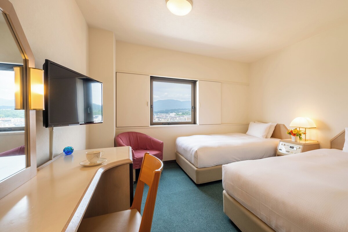 Camera d'albergo con due letti, una scrivania, una sedia, una televisione e una finestra con vista sulla città.