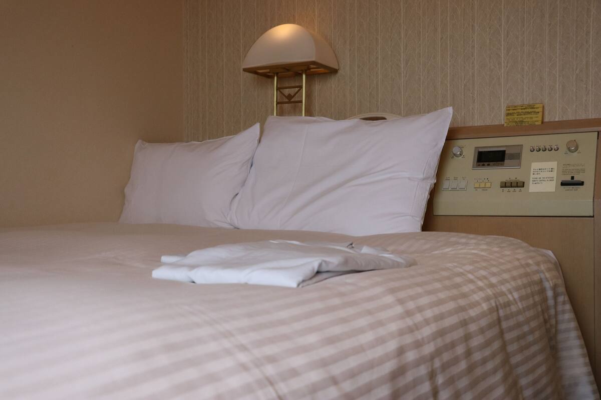 A neatly made bed with a checkered bedspread, a folded white towel, and a wall-mounted control panel with a lamp above it.