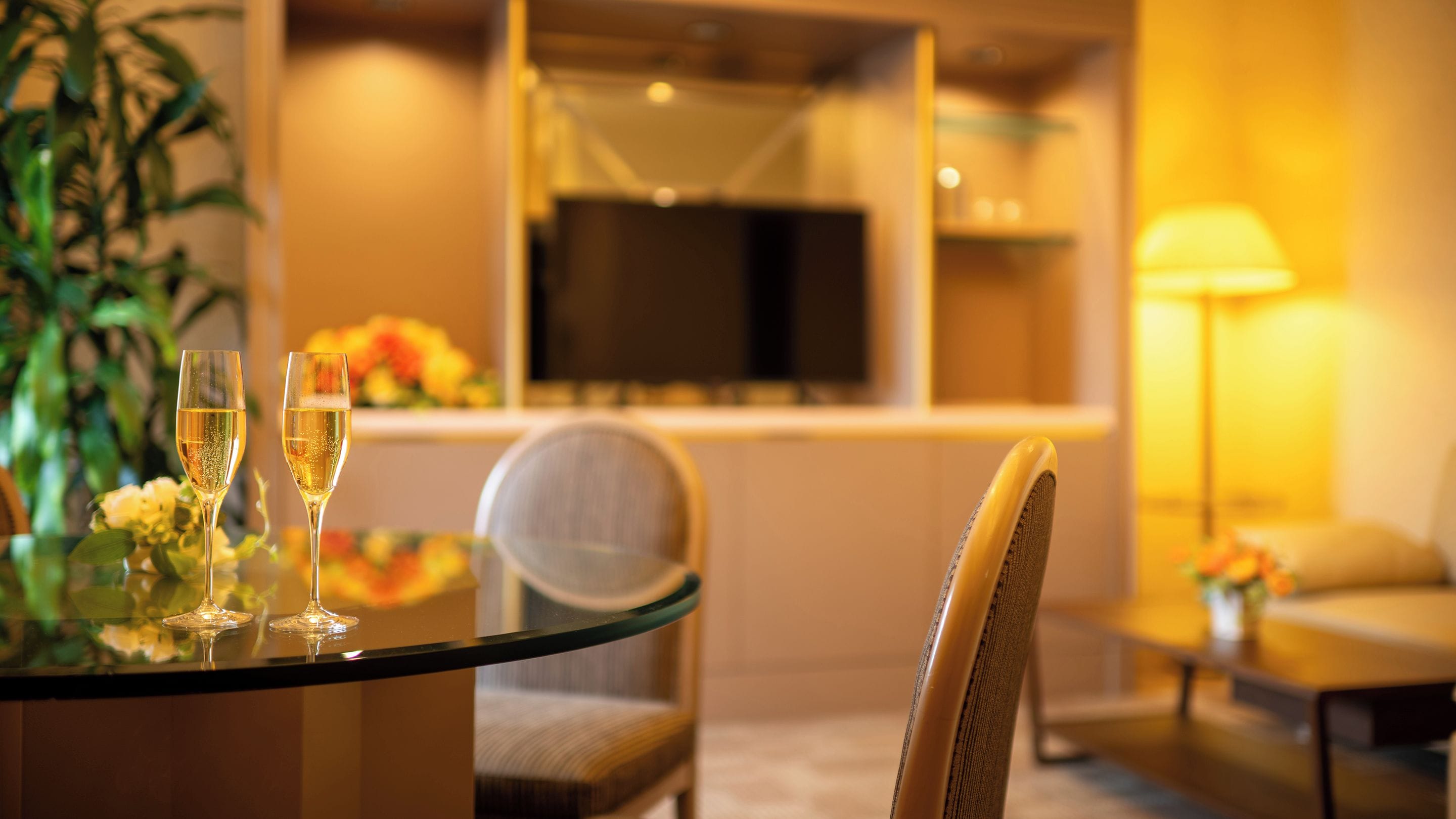 Una camera d'albergo con un tavolino in vetro, due bicchieri da champagne, un televisore e un divano.