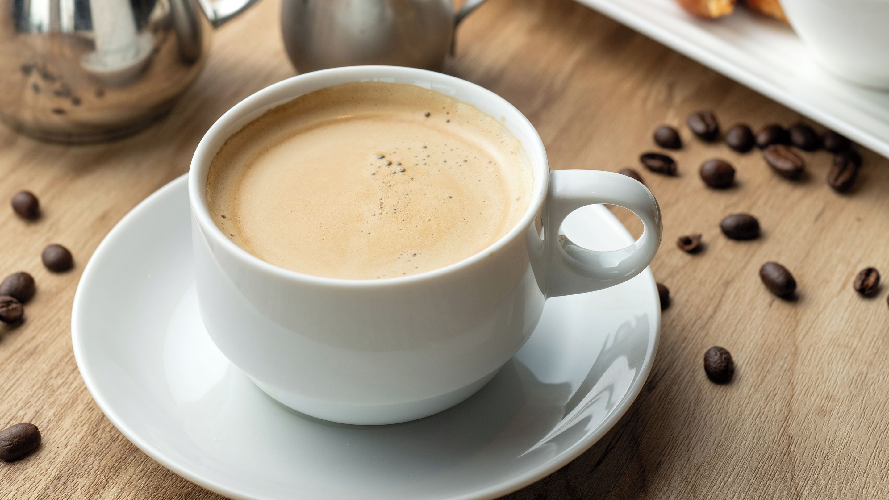 A white cup of coffee on a saucer with a handle, surrounded by coffee beans on a wooden surface.