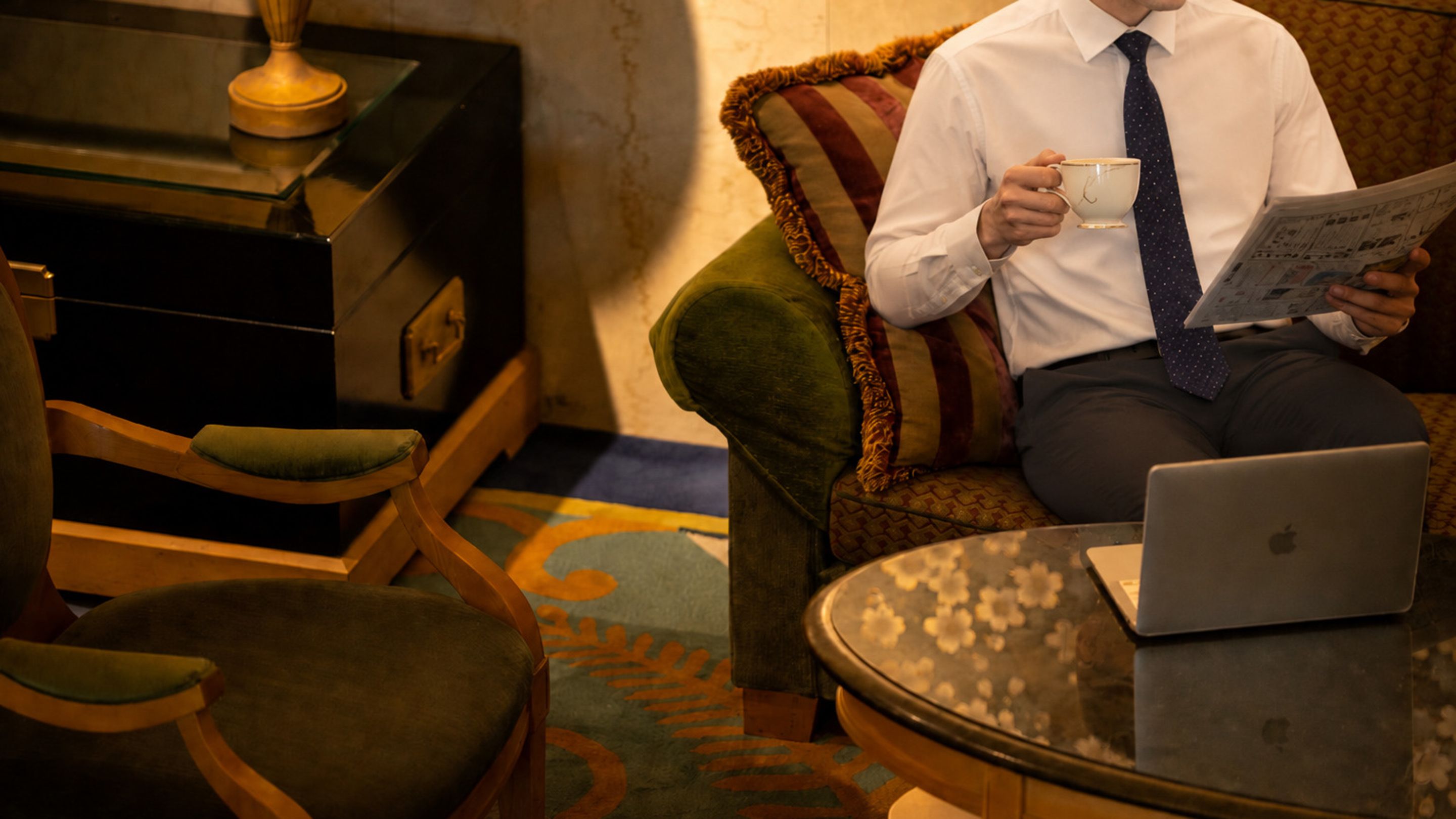 A person sitting on a sofa with a laptop and reading a newspaper.