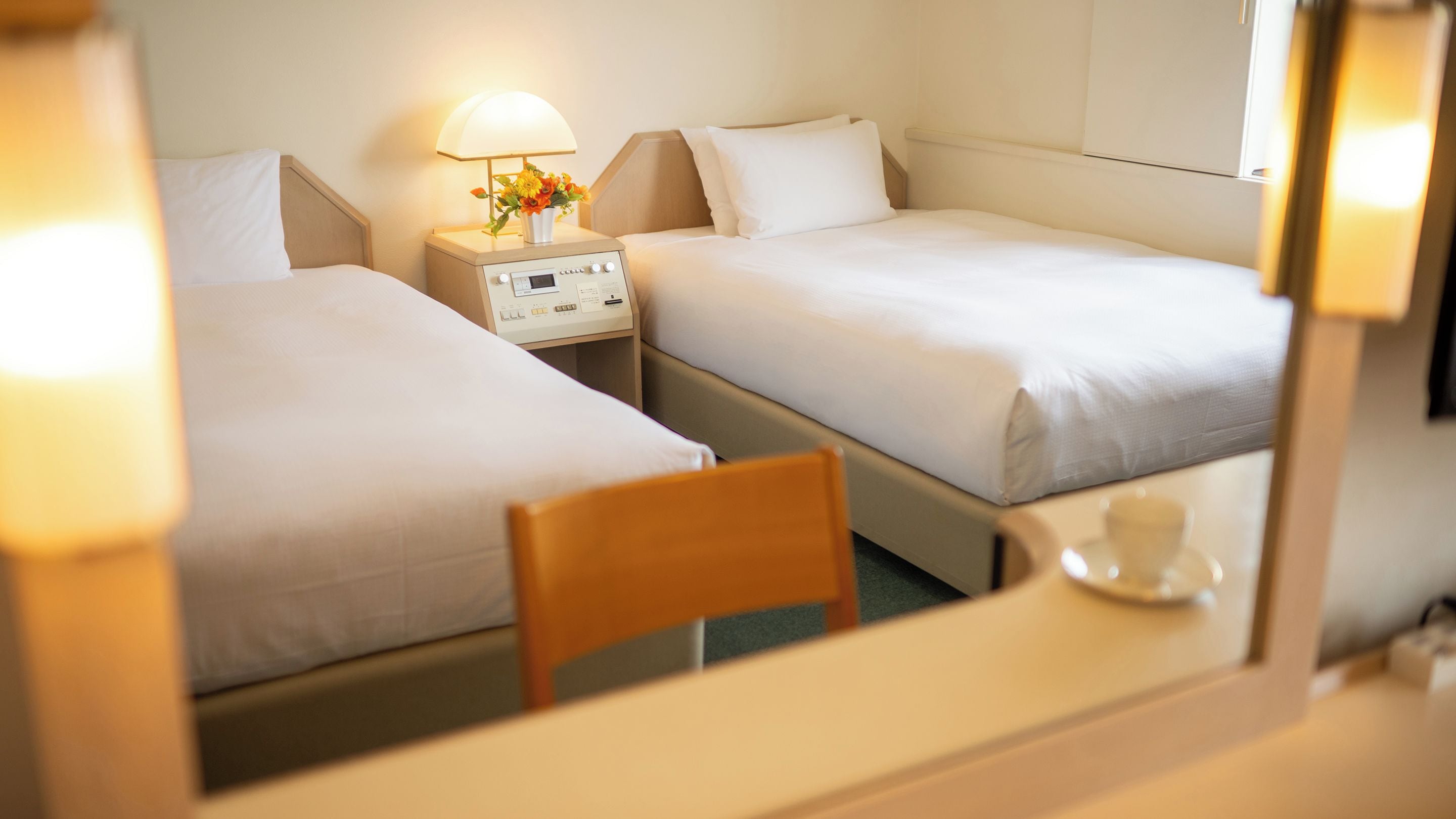 A hotel room with two beds, a bedside table with a lamp and flowers, and a chair.