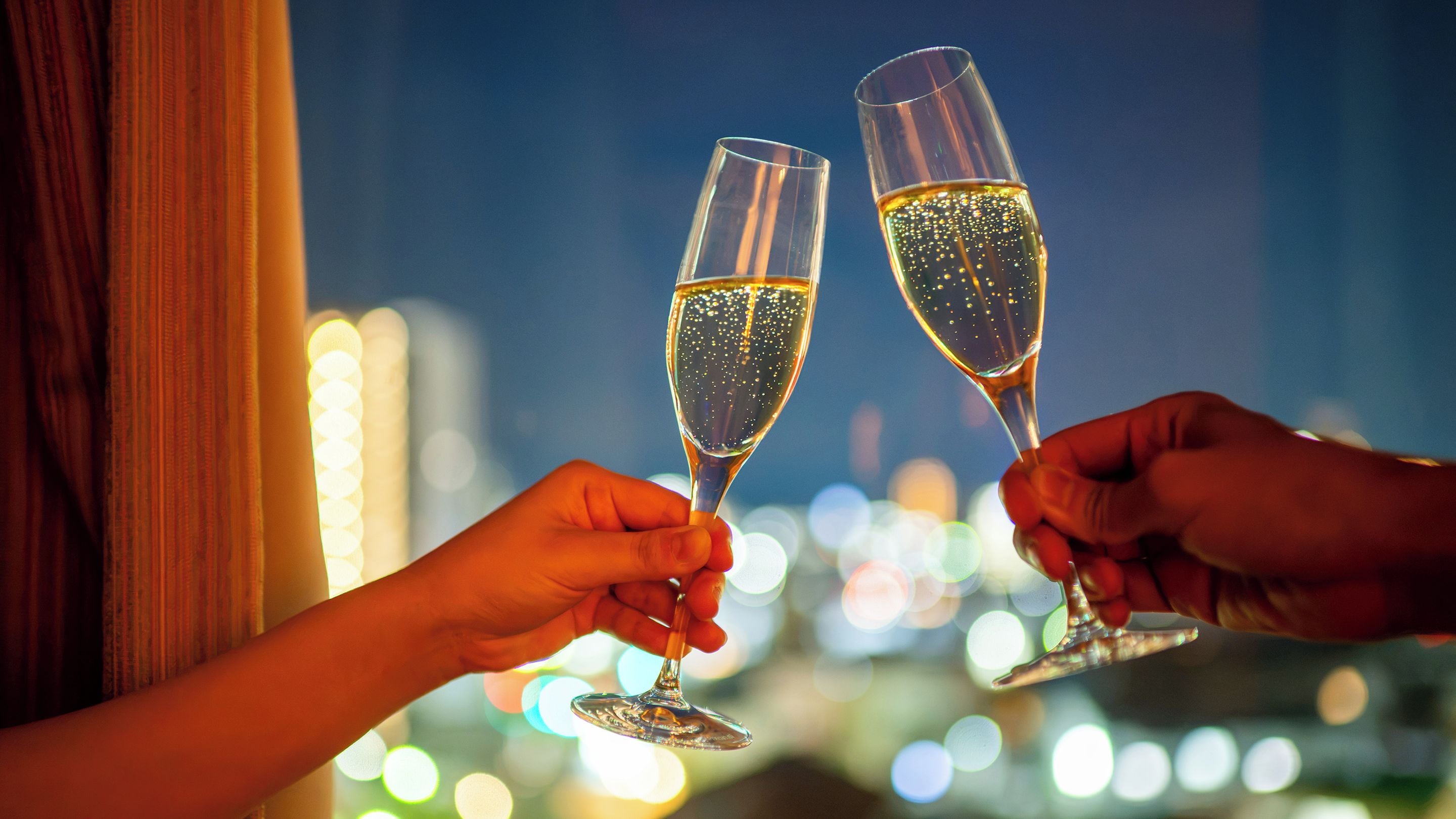 Two hands holding champagne flutes against a blurred cityscape background.