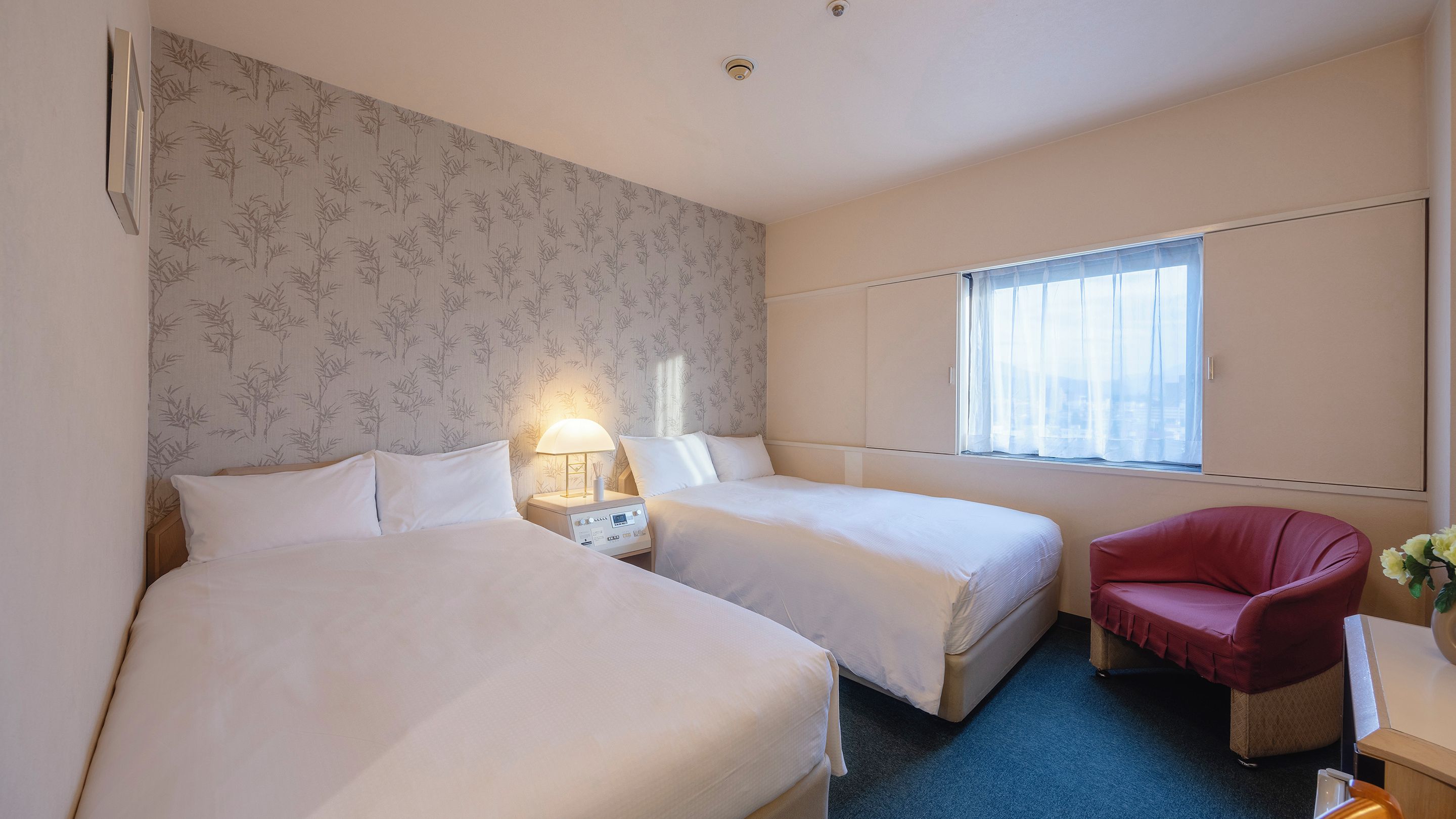 A hotel room with two beds, a red chair, a window with curtains, and a wall-mounted light fixture.