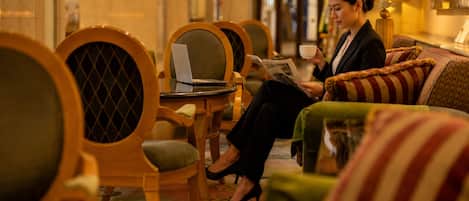Un hall de hotel con disposición de asientos, una mesa con una computadora portátil y una persona leyendo una revista.