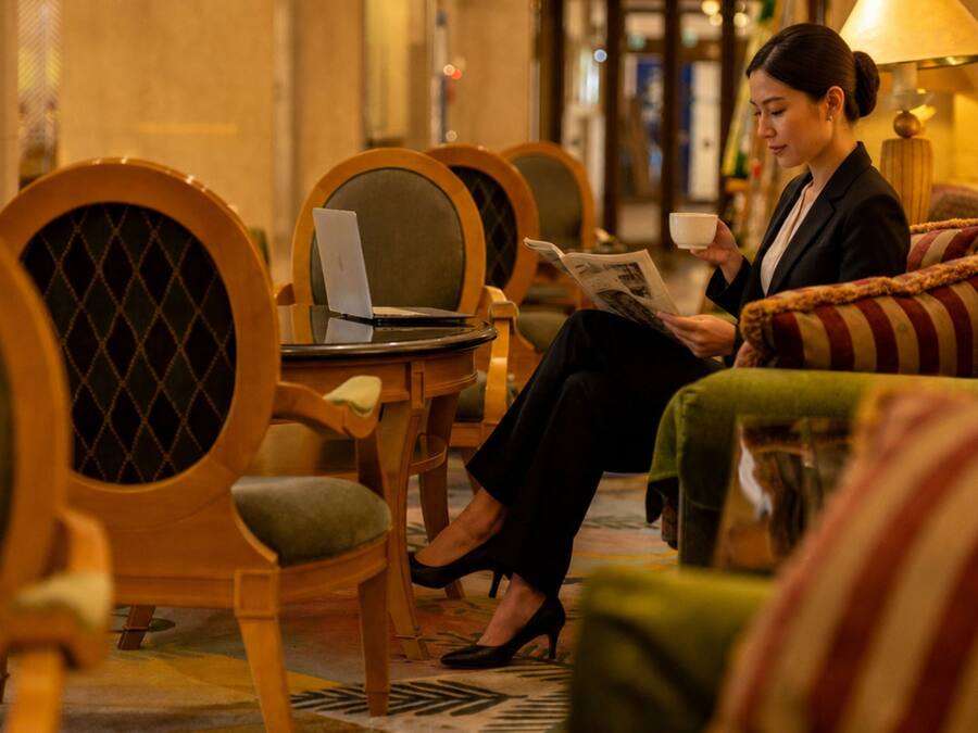 A hotel lobby with seating arrangements, a table with a laptop, and a person reading a magazine.