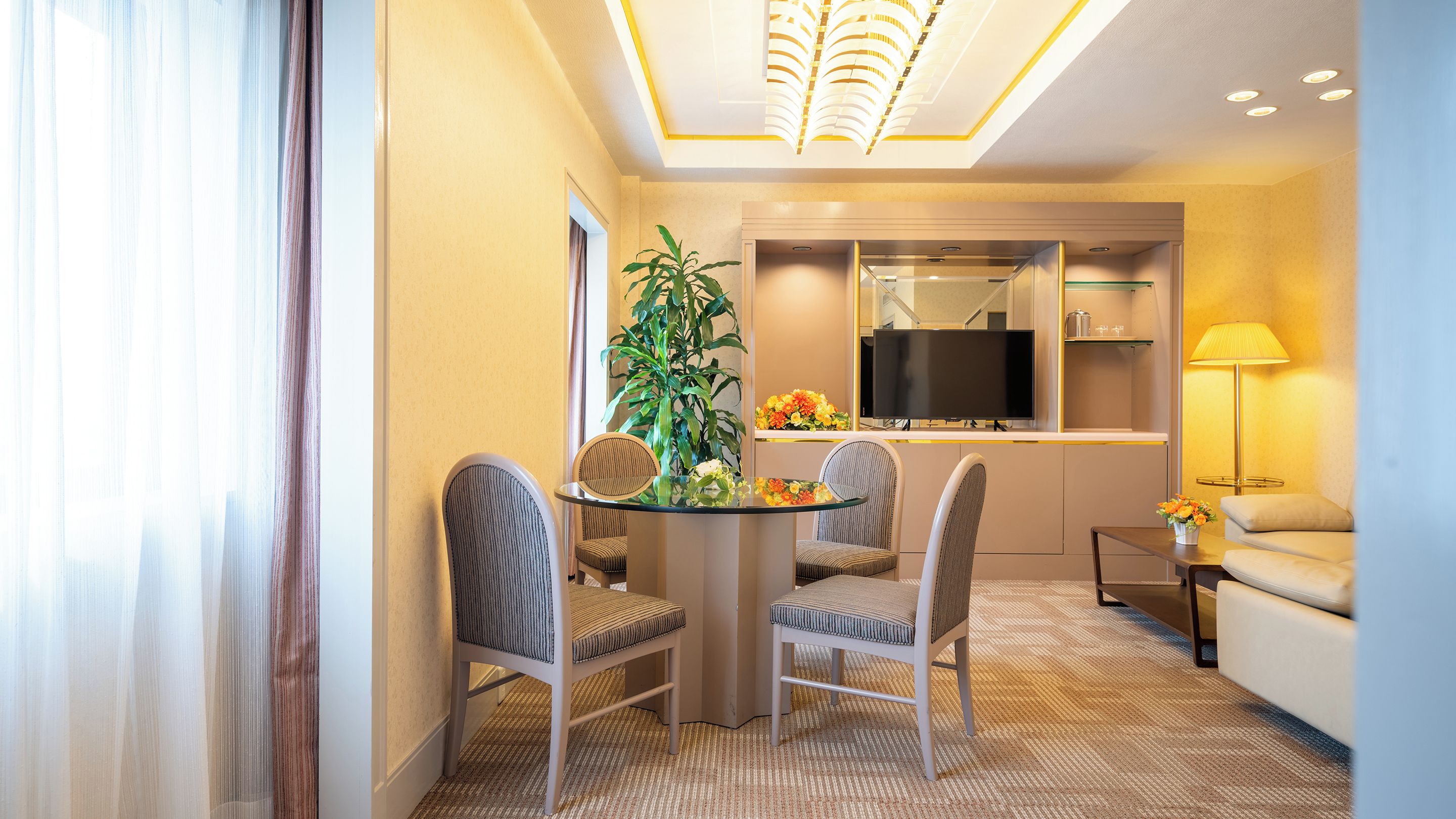 A hotel room with a dining area featuring a round table, two chairs, a potted plant, and a flat-screen TV.