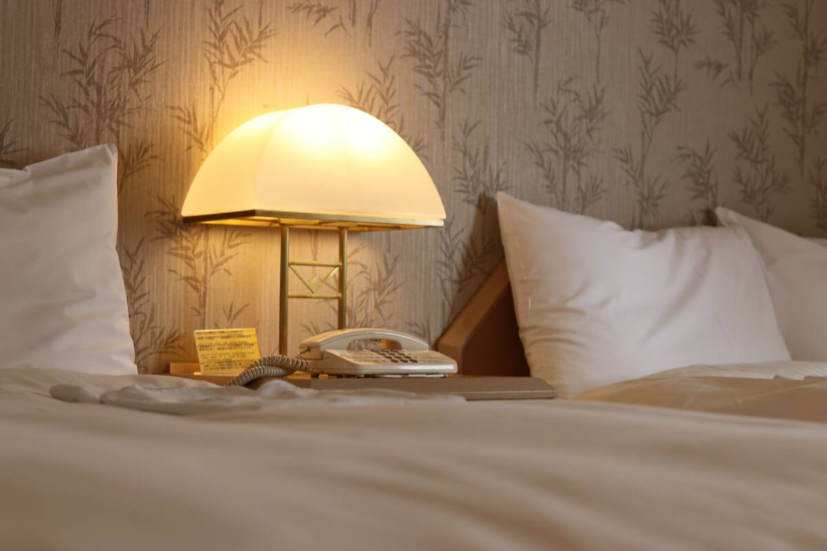 A neatly made bed with white linens, a bedside lamp, a telephone, and a notepad on a wooden nightstand.