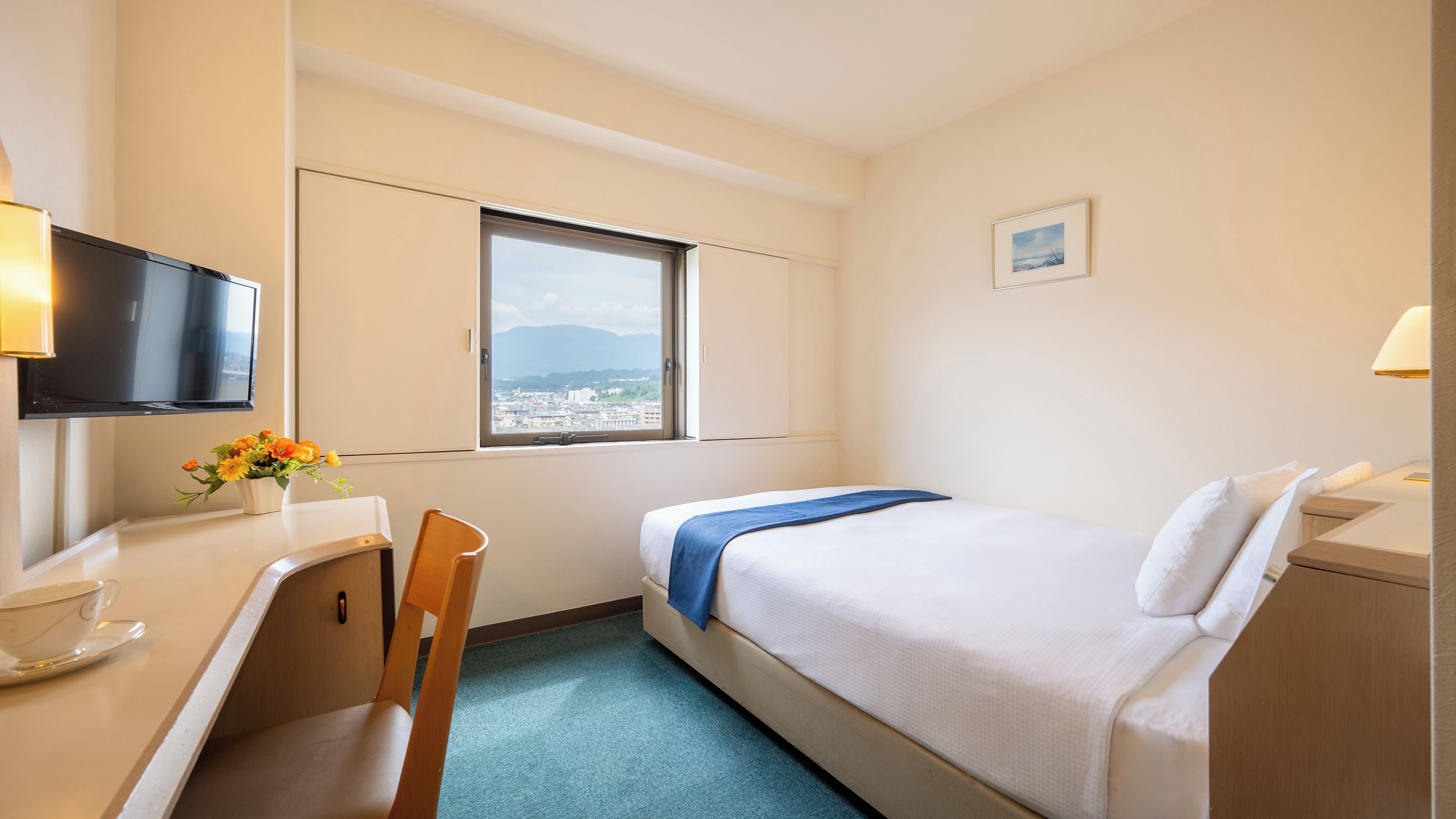 A hotel room with a bed, a desk, a chair, a TV, a window with a view, and a small picture on the wall.