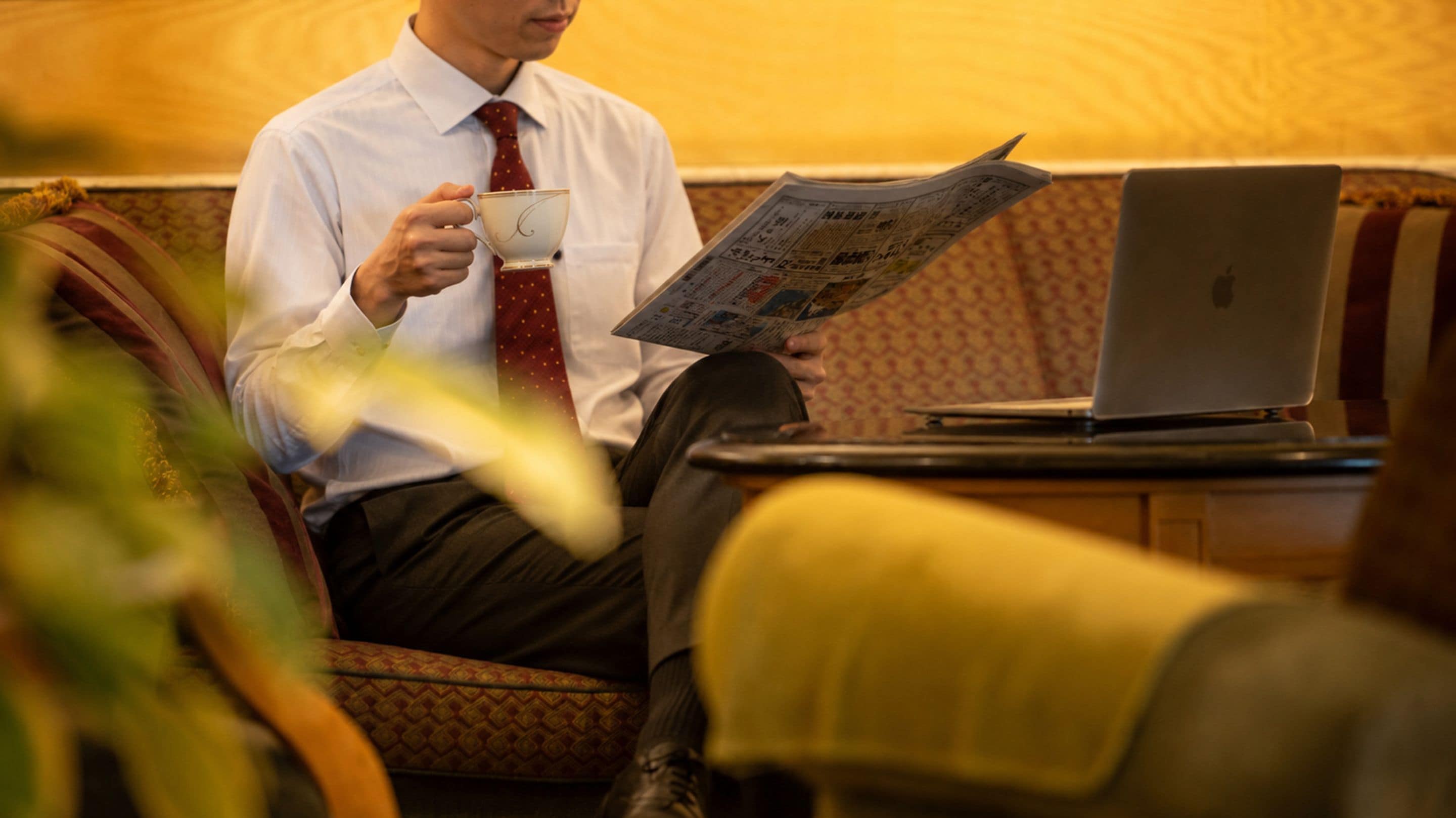 A person sitting on a patterned sofa with a laptop on a table beside them, holding a newspaper.
