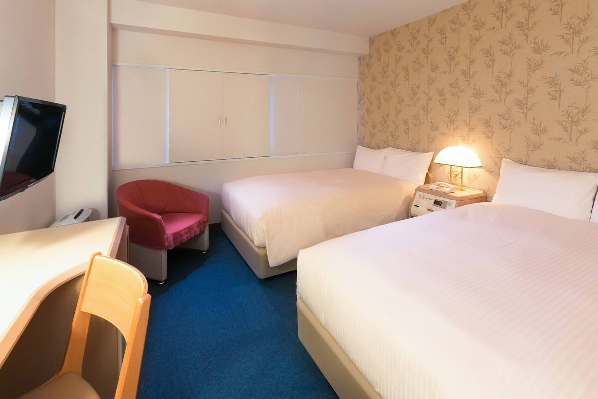 A hotel room with two beds, a desk, a chair, a TV, and a nightstand with a lamp.