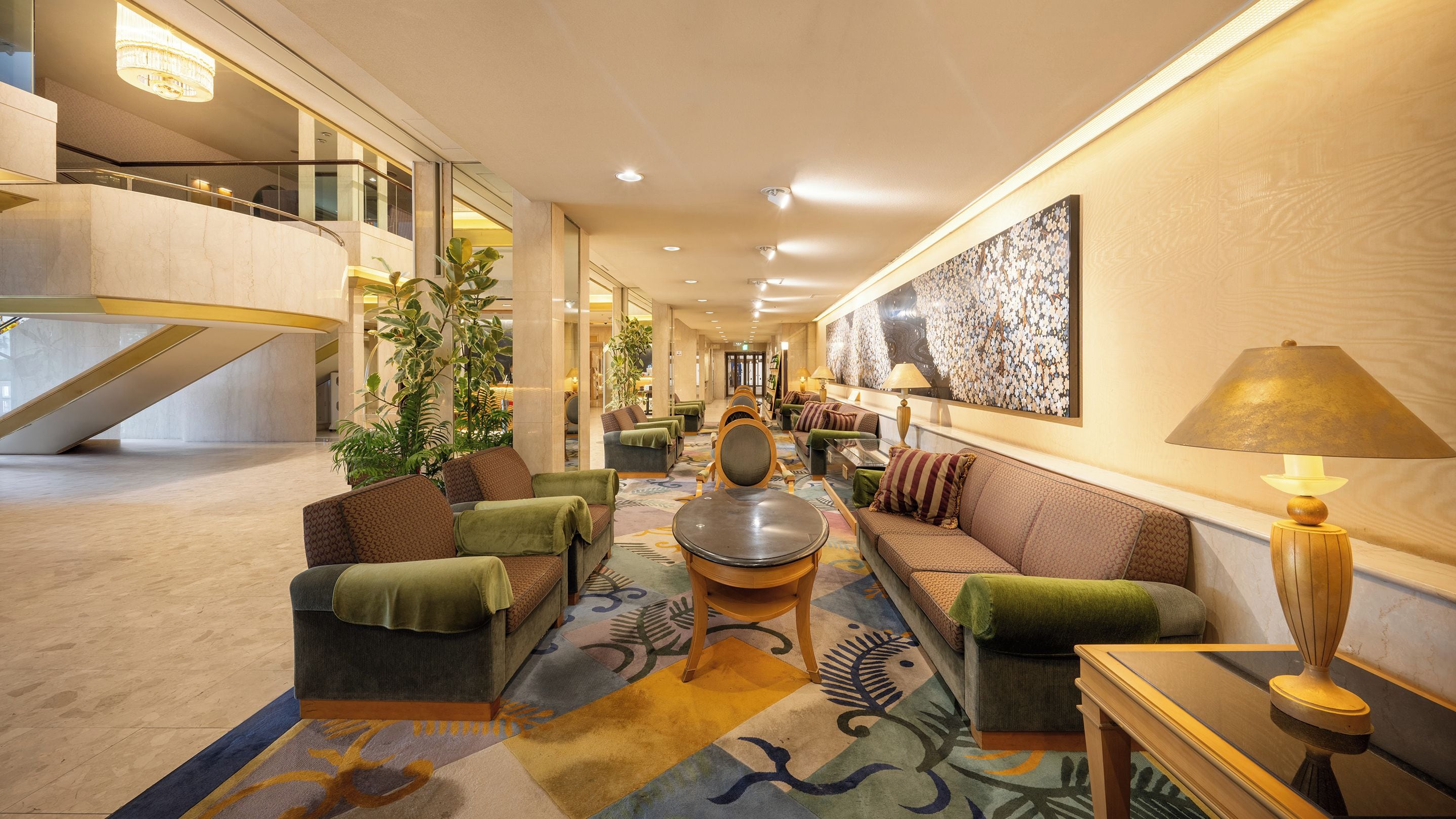 A spacious lobby with a high ceiling, a staircase, seating area with couches and chairs, a coffee table, and a large abstract painting on the wall.