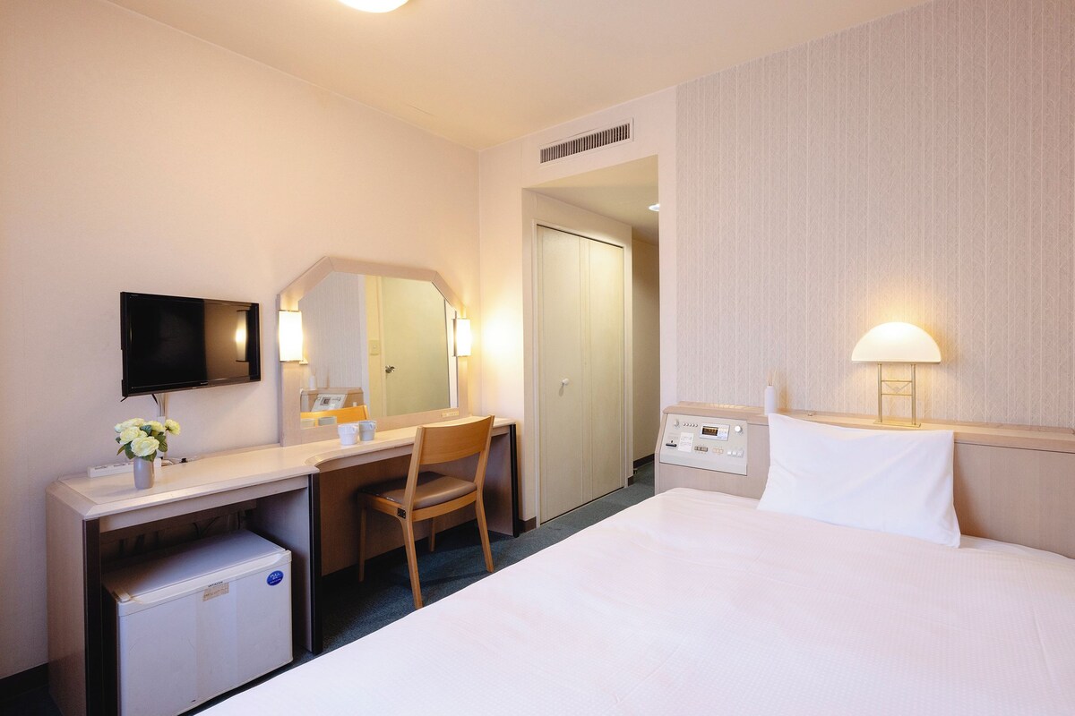 Una camera d'albergo con un letto, una scrivania, una sedia, una televisione e un comodino.