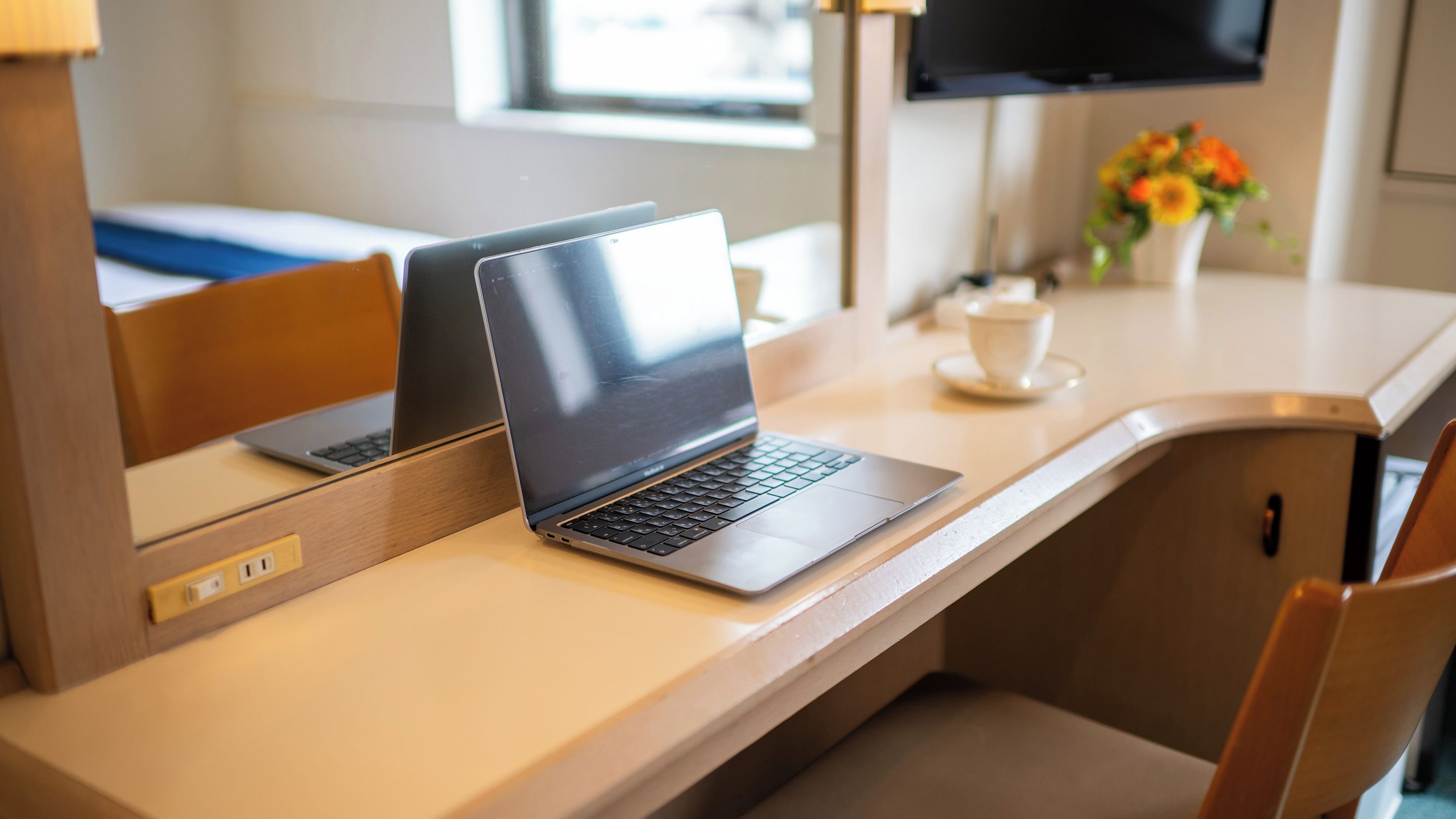 A laptop on a wooden desk with a cup and saucer, a television, and a flower arrangement in a hotel room.