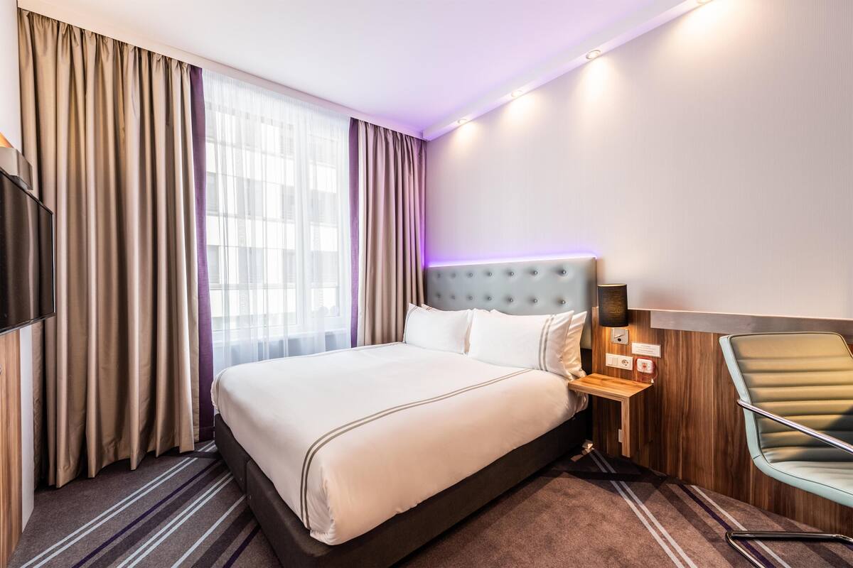 Una habitación de hotel con una cama grande, televisión, mesita de noche, lámpara y una silla.