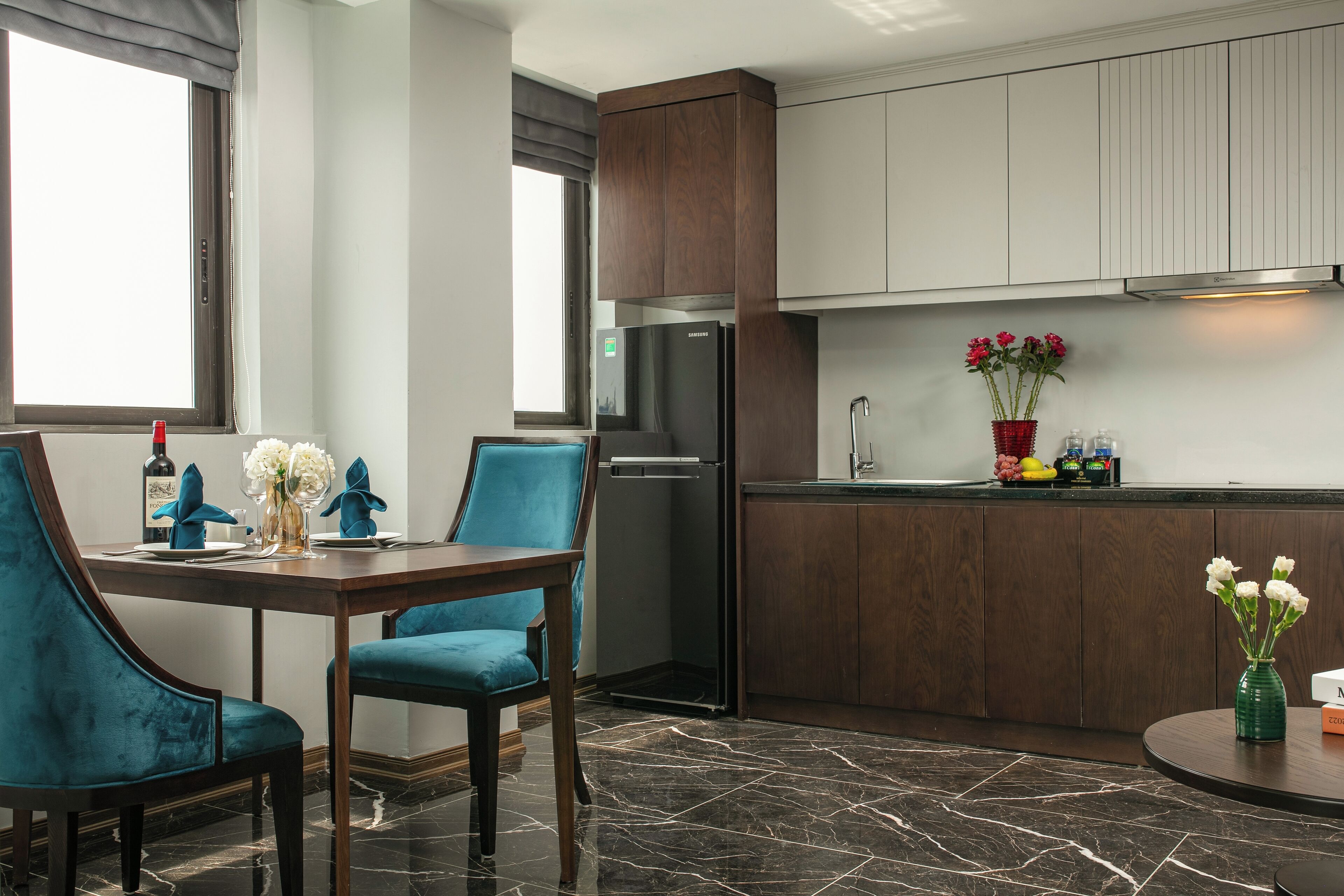 A modern kitchen with dark wood cabinets, a stainless steel refrigerator, and a dining area with teal chairs and a marble floor.