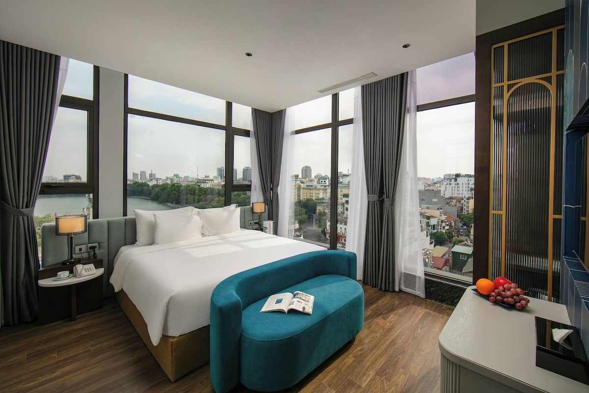 Une chambre d’hôtel moderne avec un grand lit, un canapé bleu, une petite table avec une lampe et une vue sur la ville grâce à de grandes fenêtres.