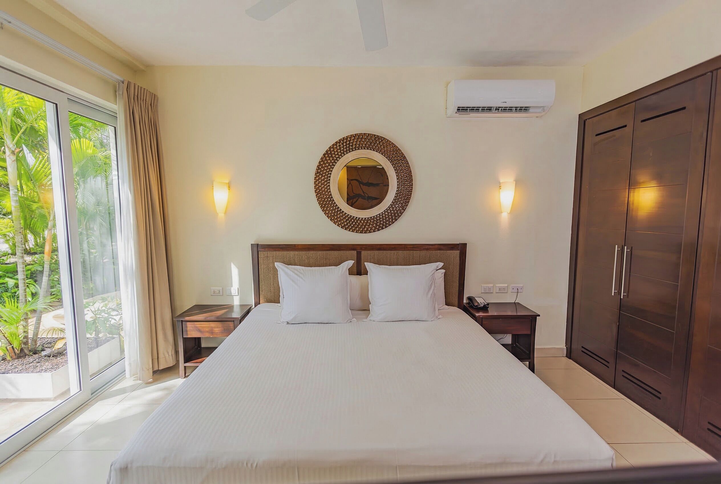 A hotel room with a bed, bedside tables, a wardrobe, and a view of greenery.