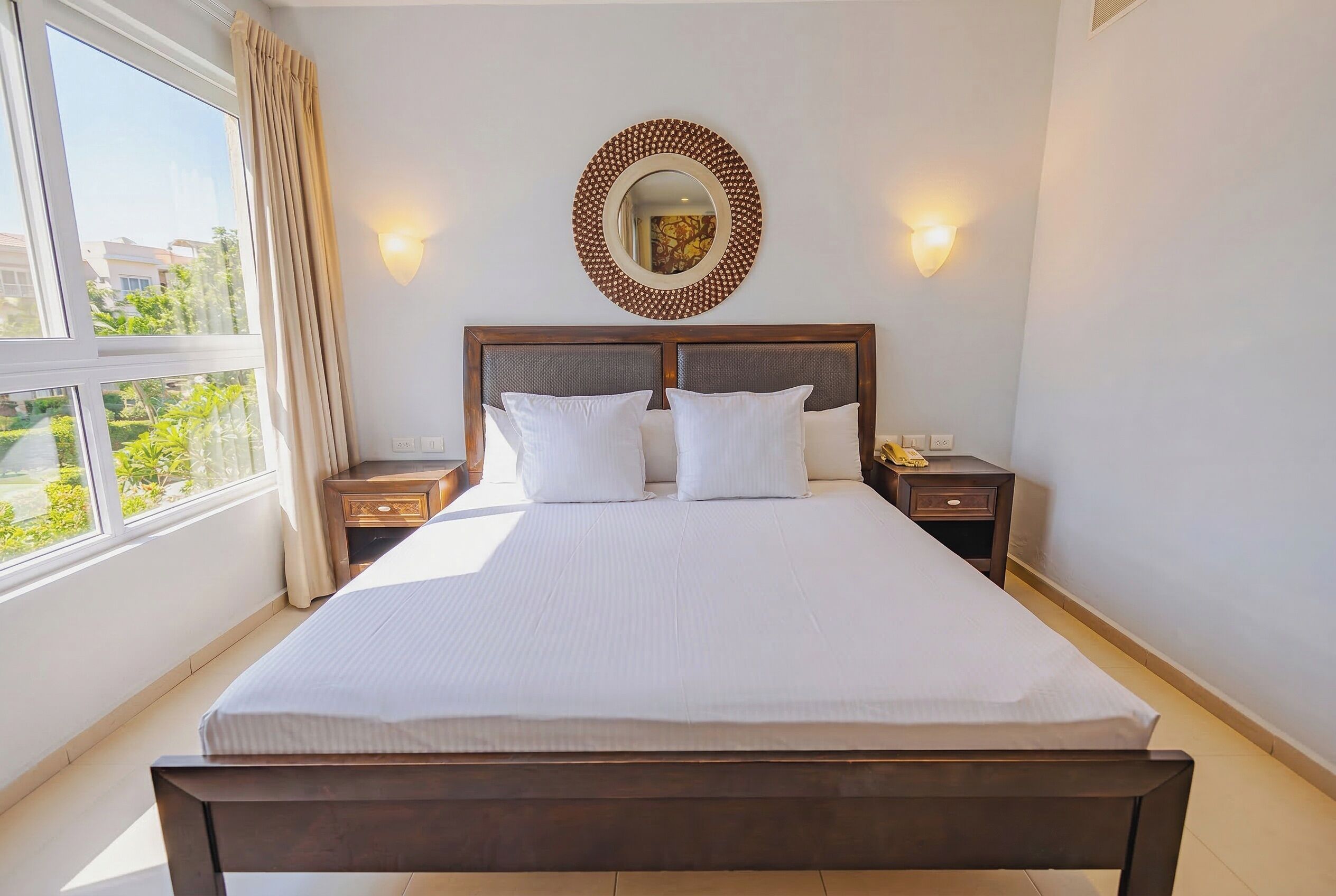 A hotel room with a large bed, two bedside tables, a round mirror, and a view of greenery outside.