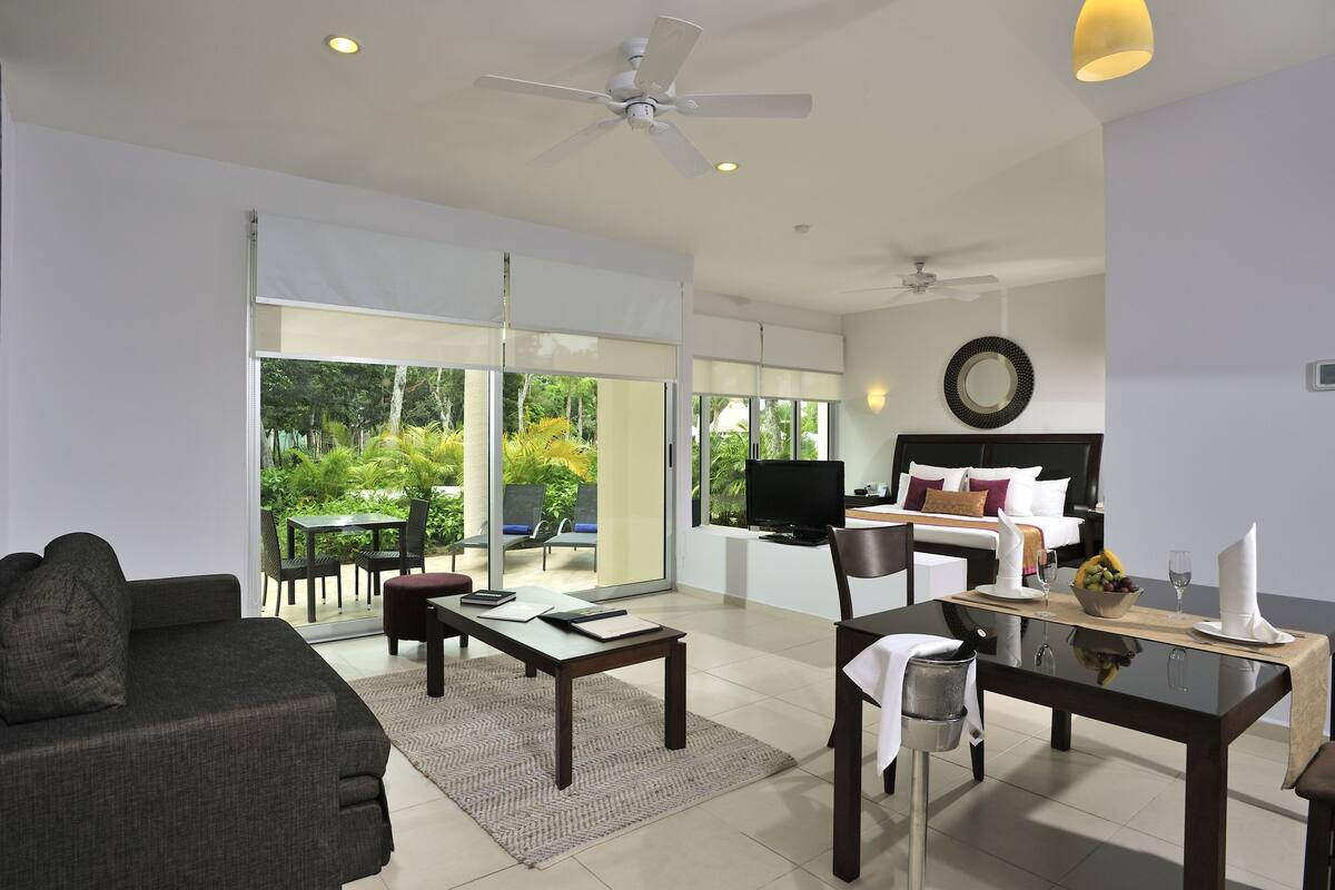 A modern living room with a sofa, coffee table, and dining table. There is a TV, a bed with a headboard, and a view of greenery outside.