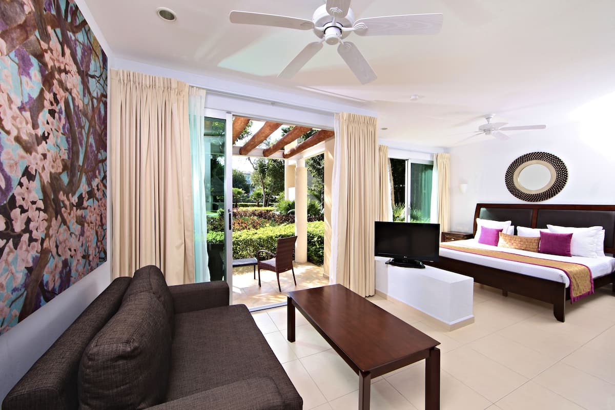 A hotel room with a sofa, a coffee table, a bed with a TV, and a balcony with curtains.