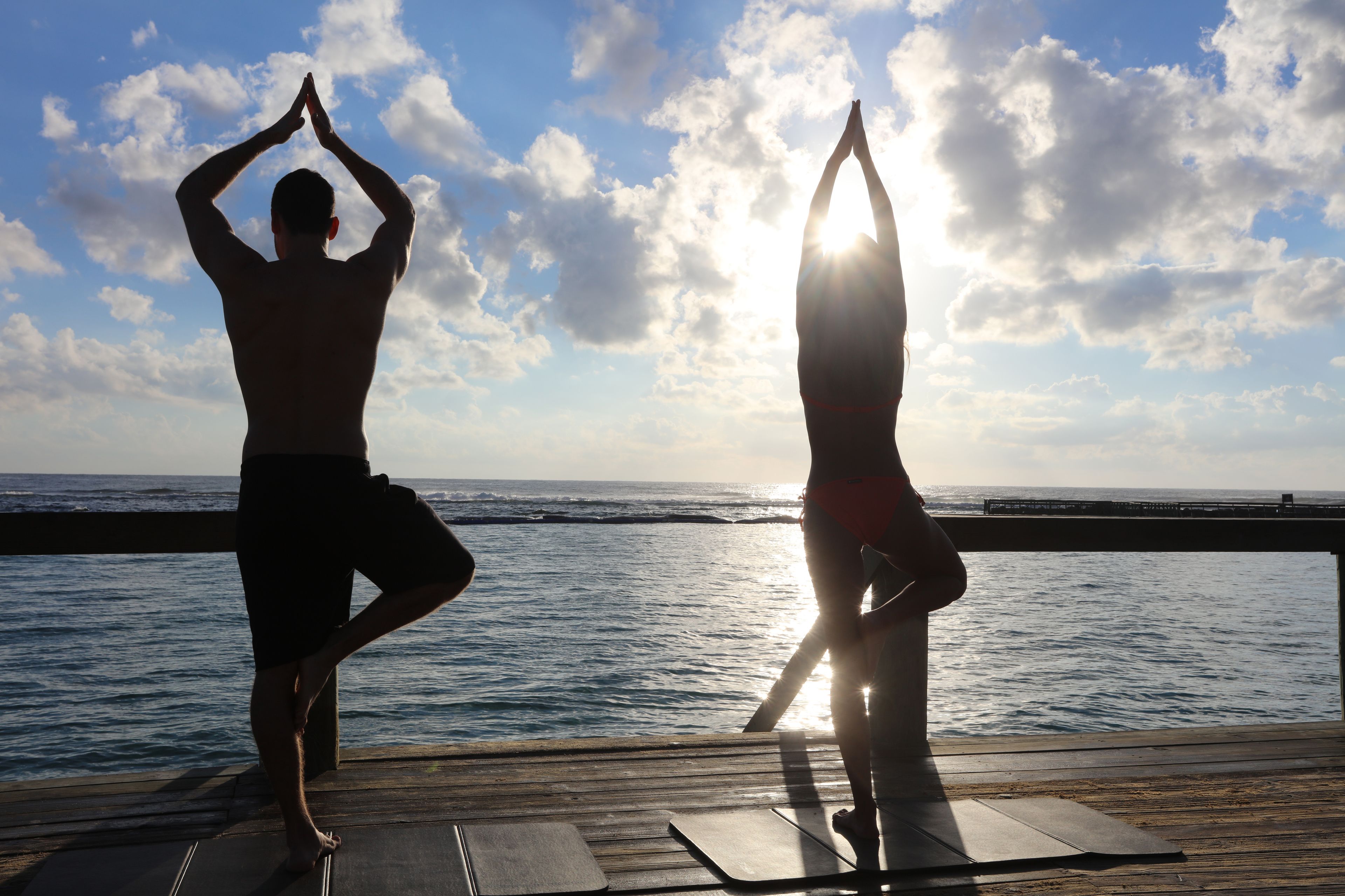 Two people practicing yoga on a wooden deck with a view of the ocean and a cloudy sky.