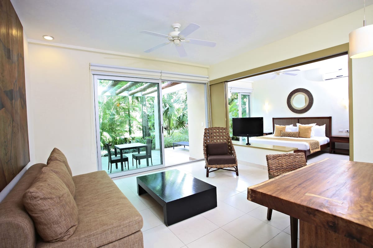A modern hotel room with a sofa, a wooden coffee table, a dining table, a wicker chair, a bed with a headboard, and a large window offering a view of the outdoors.