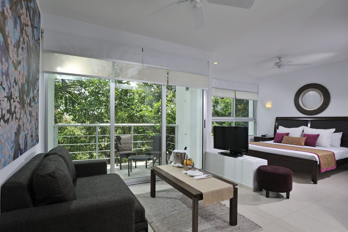 A modern hotel room with a large sliding glass door leading to a balcony, a bed with a TV, a sofa, and a small table.