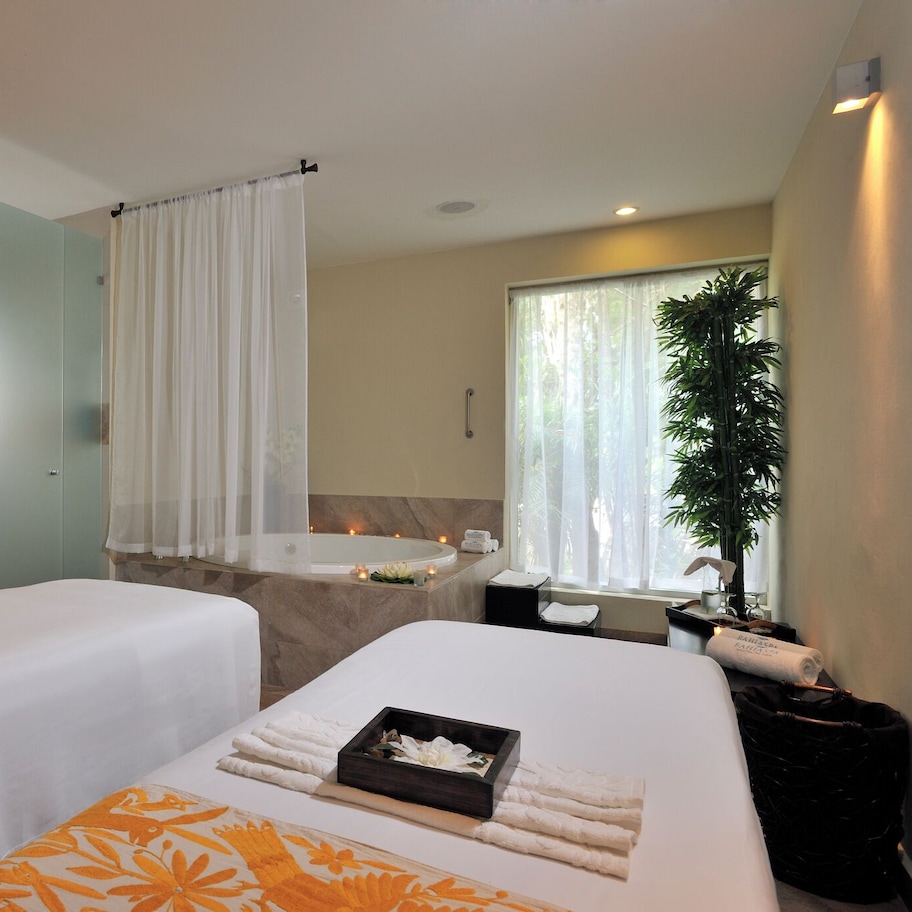 A hotel room with two beds, a bathroom with a bathtub, a large window with a plant, and a mirror with lights.