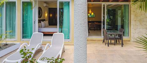 A hotel room with a bed, a sitting area with a table and chairs, and a balcony with lounge chairs.