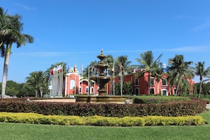 Un edificio rojo con una fuente en primer plano, rodeado de palmeras y setos cuidados.