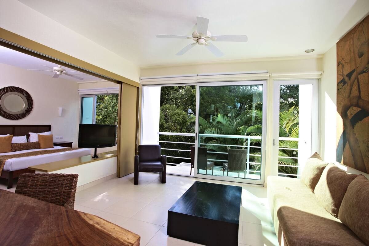 A modern hotel room with a bed, a flat-screen TV, a sofa, a coffee table, and a balcony with glass doors.