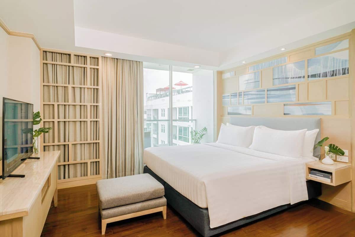 Une chambre d’hôtel moderne avec un grand lit, un bureau avec une télévision, un petit banc et une vue sur la ville grâce à de grandes fenêtres.