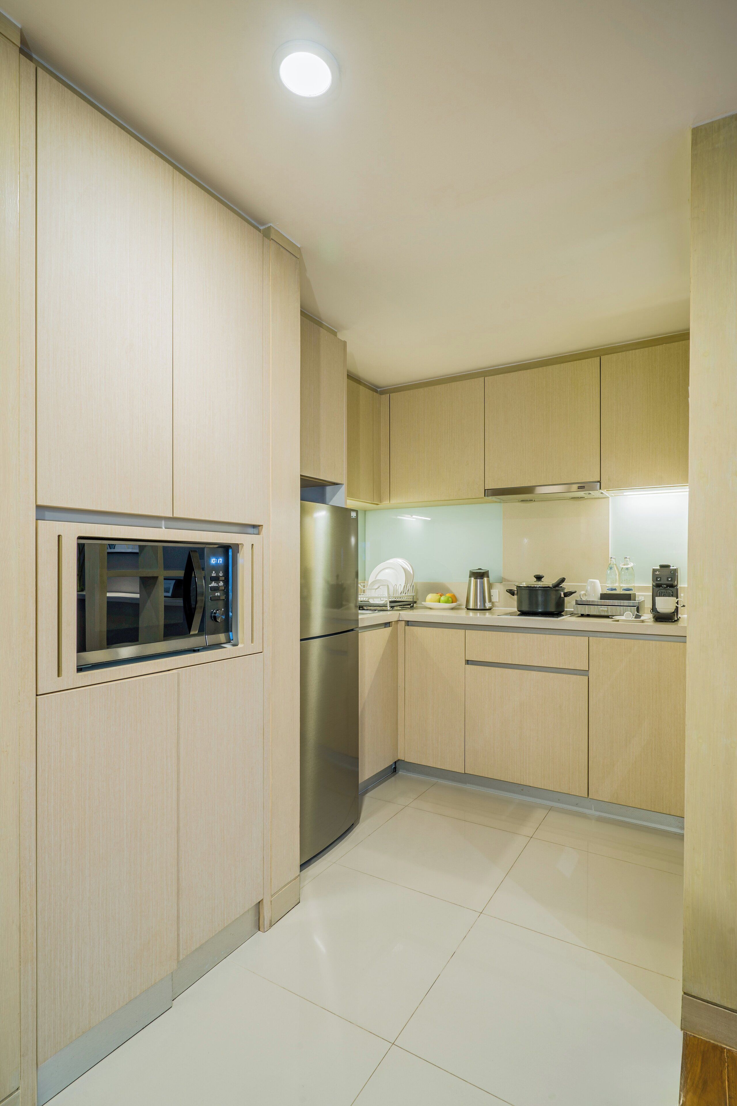 A modern kitchen with light wood cabinets, a built-in microwave, stainless steel appliances, and a clean tiled floor.