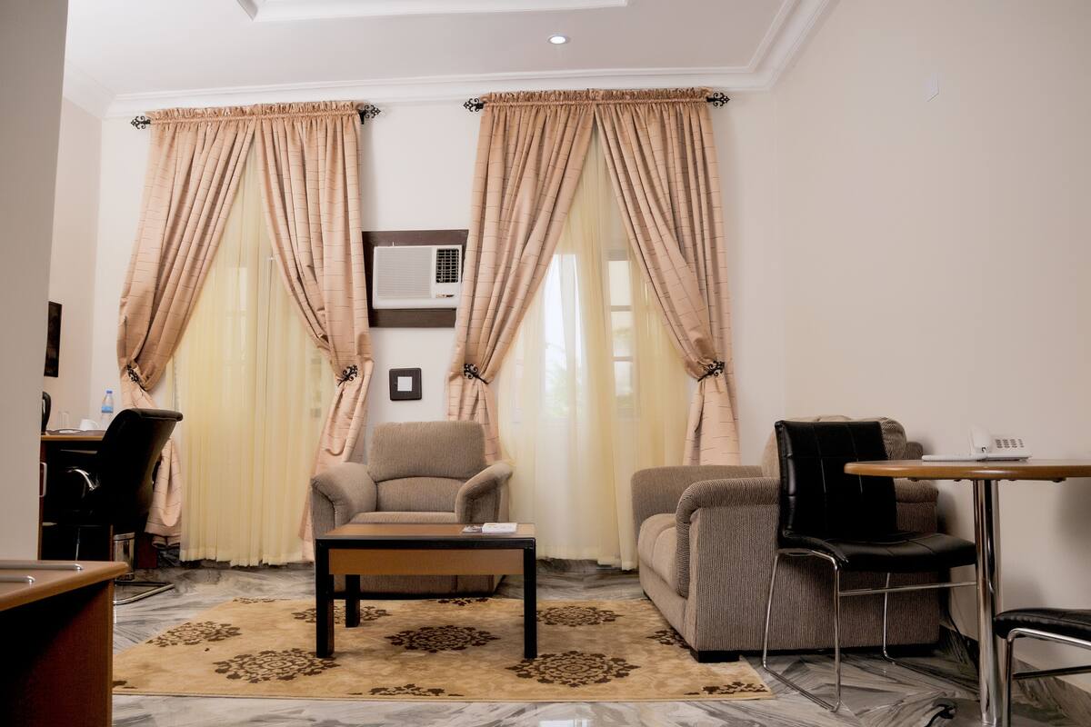 A living room with a sofa, armchair, and a small table. There is a window with curtains, a wall-mounted air conditioner, and a picture frame.
