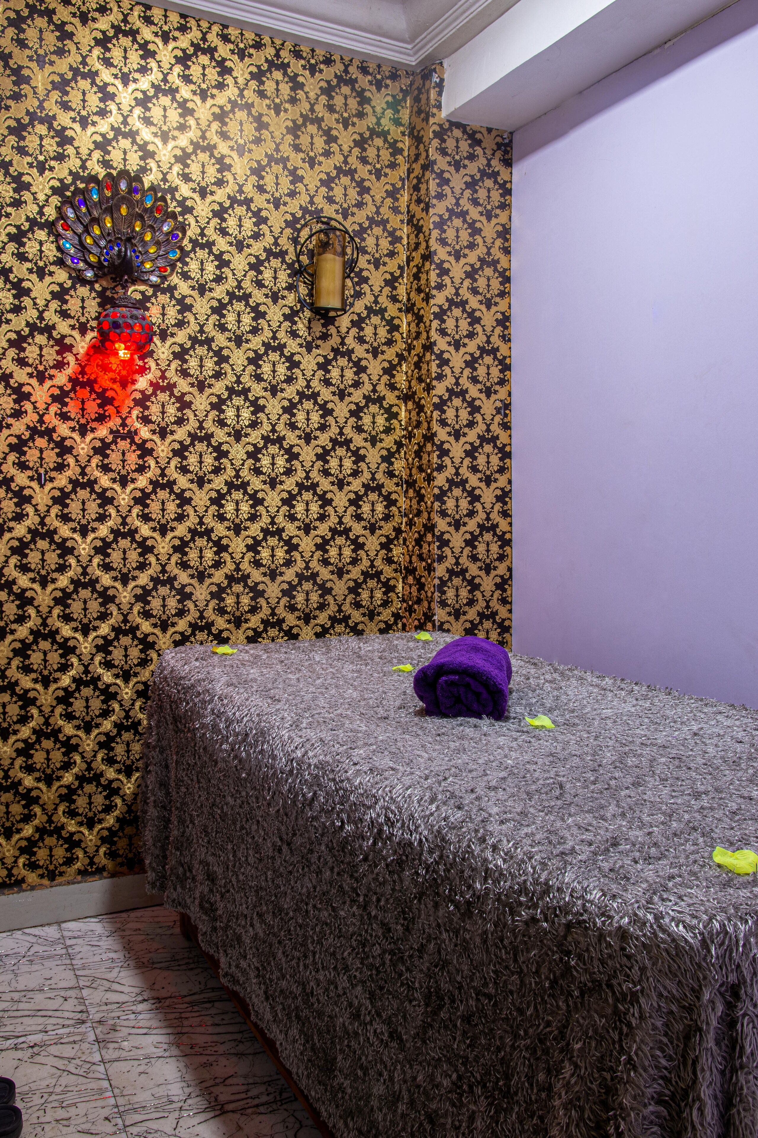 A room with a patterned wallpaper, a bed with a grey textured cover, and a peacock-themed wall decoration.