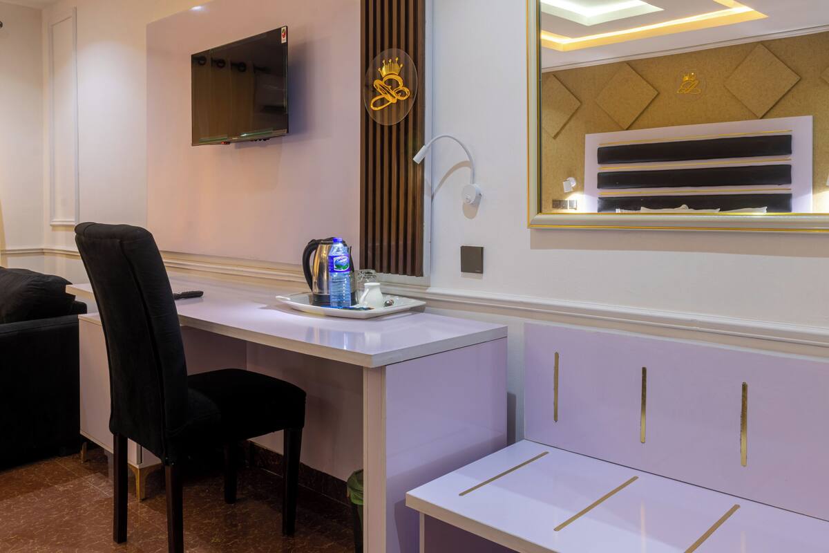 A modern hotel room with a white desk, a black chair, a flat-screen TV mounted on the wall, and a decorative wall panel with a crown symbol.