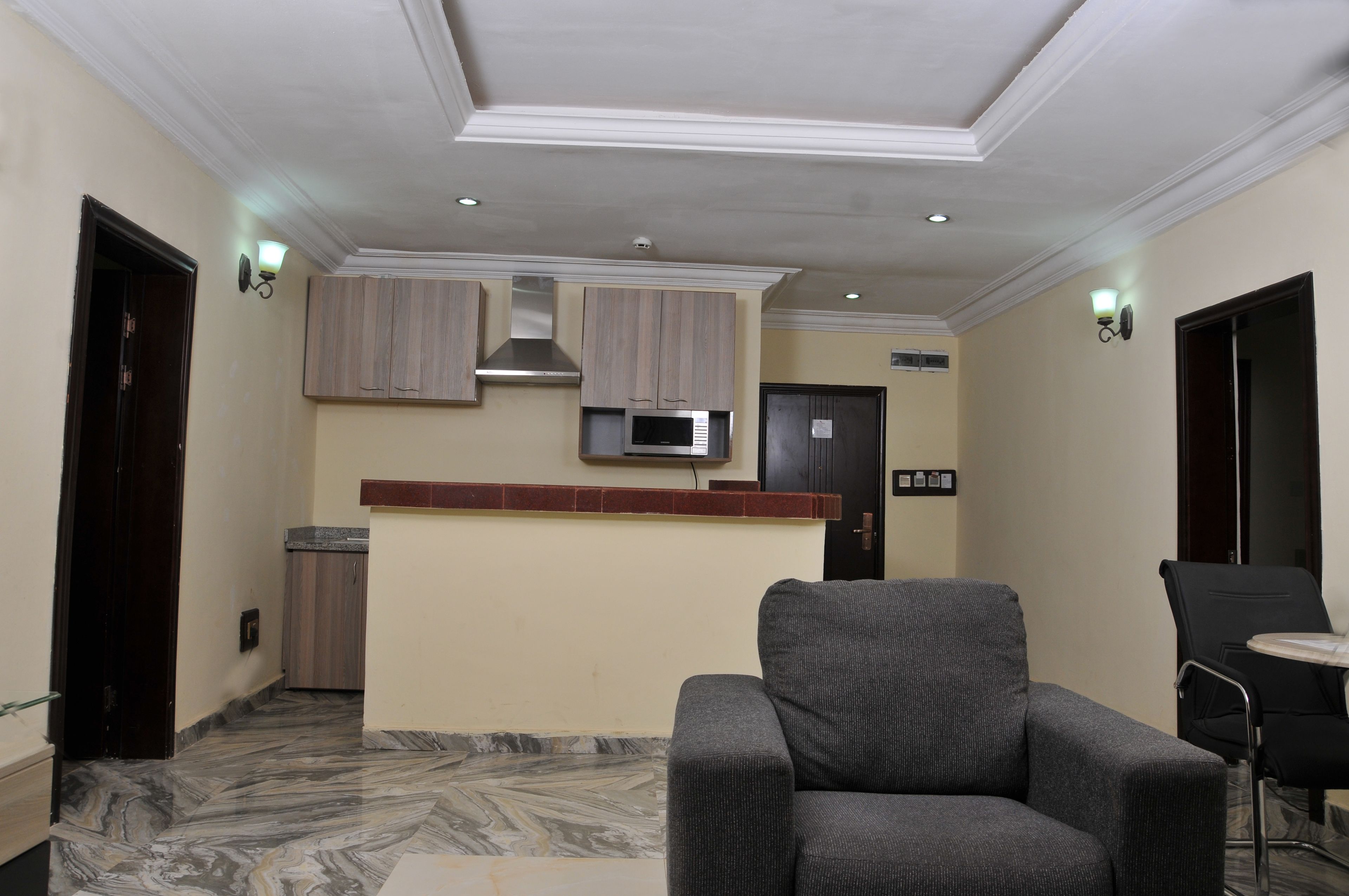 A modern kitchen with wooden cabinets, a stainless steel range hood, and a microwave. There is a dining area with a grey upholstered chair and a small table.