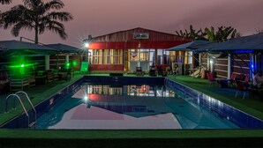 A pool area with a red building labeled "JOY HALL" in the background, surrounded by outdoor seating and a palm tree. - Hotel De Bently (Abuja)
