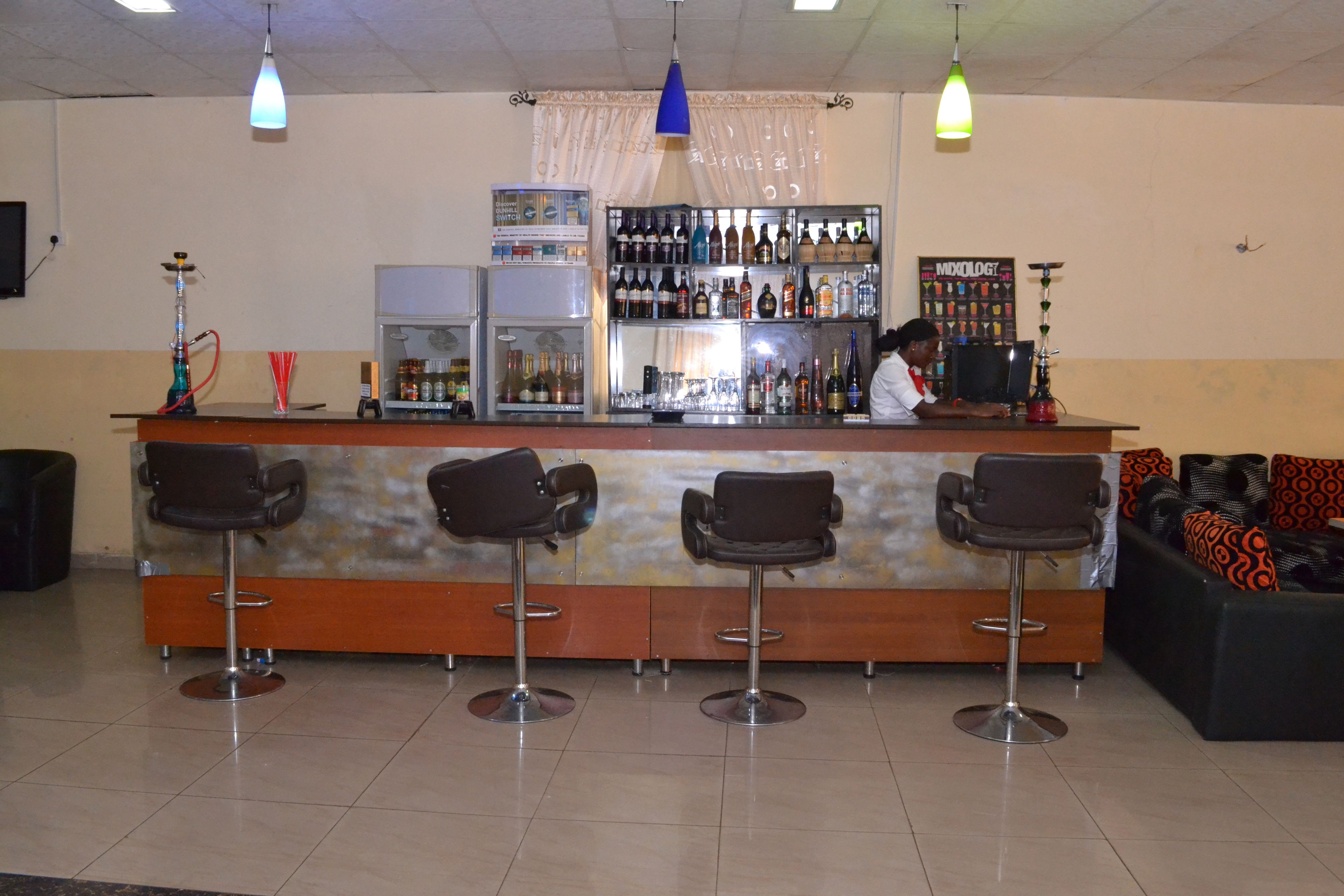 A bar area with a counter, multiple stools, a refrigerator, shelves with bottles, and a sofa with patterned cushions.