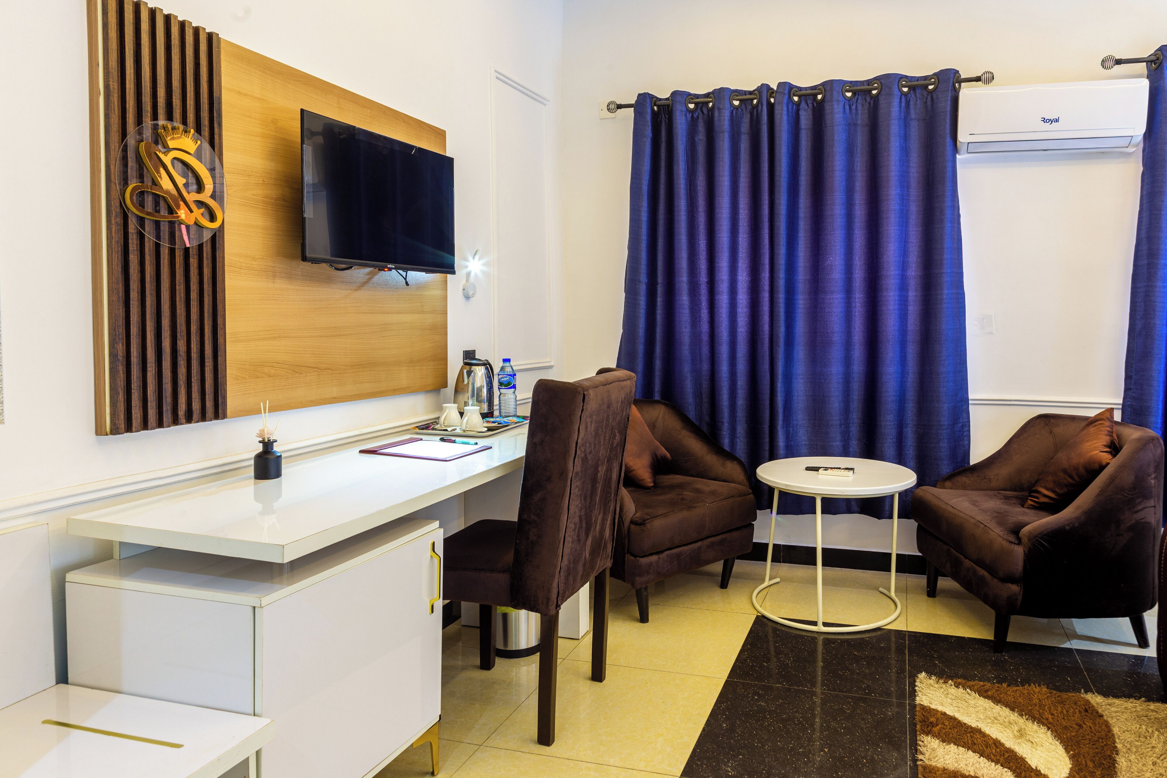 Quarto de hotel com uma escrivaninha branca, duas poltronas marrons, uma pequena mesa redonda, uma TV de tela plana presa a um painel de madeira, uma cortina azul e um aparelho de ar condicionado.