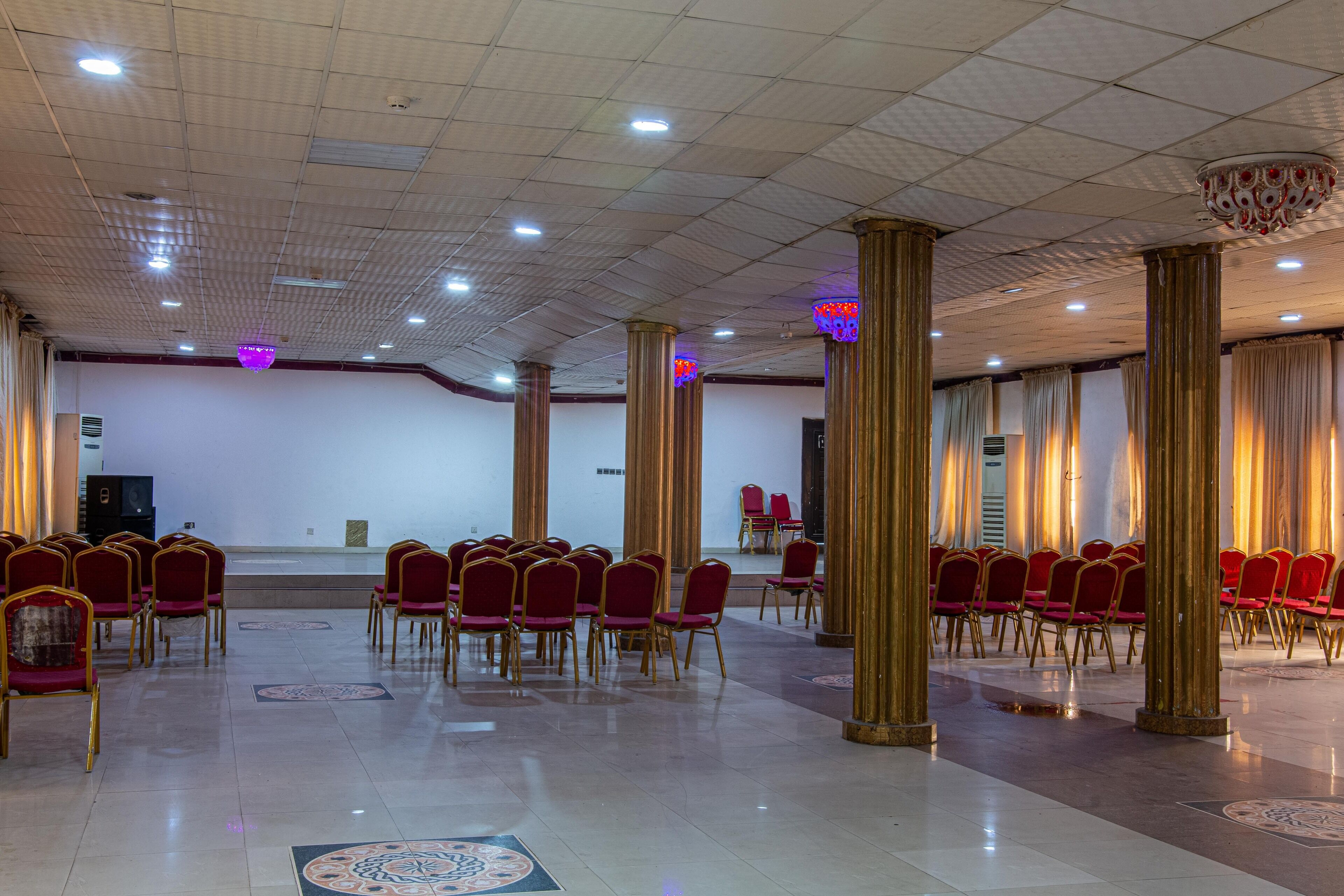 A spacious hall with rows of red chairs and gold columns, a stage, and patterned floor tiles.