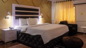 A hotel room with a large bed, a bedside table with a phone, a chair, a small table, and a curtain. - Hotel De Bently (Abuja)